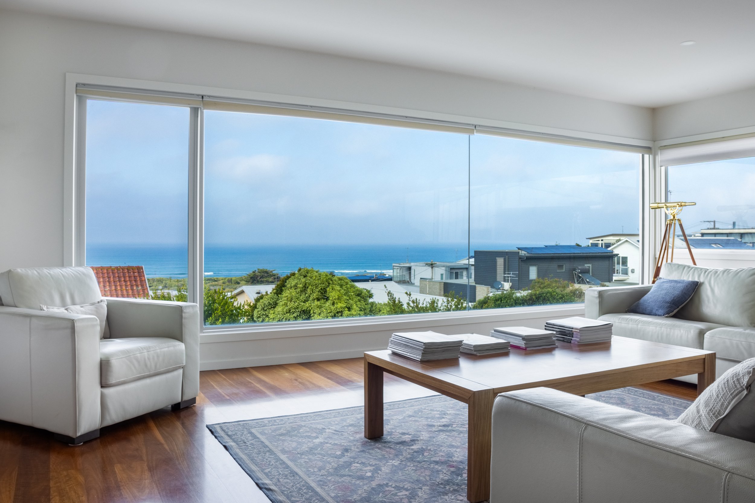 Living room with white sofa, wooden coffee table with stacks of magazines, large window with ocean view, telescope, and small rind tap on ceiling.
