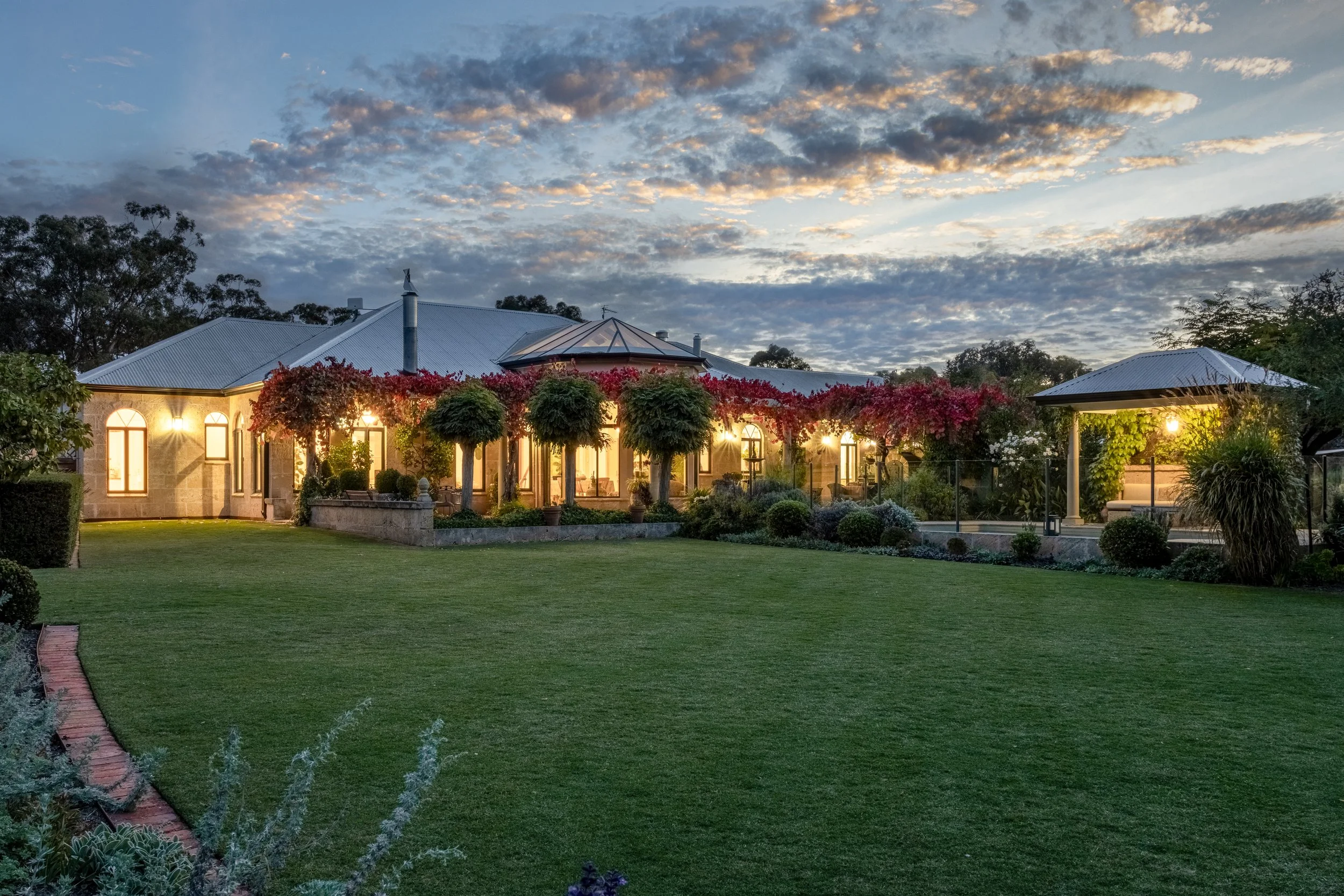 A house at dusk with illuminated windows and outdoor lights, surrounded by a well-maintained lawn, trees, and flowering plants, under a cloudy sky.