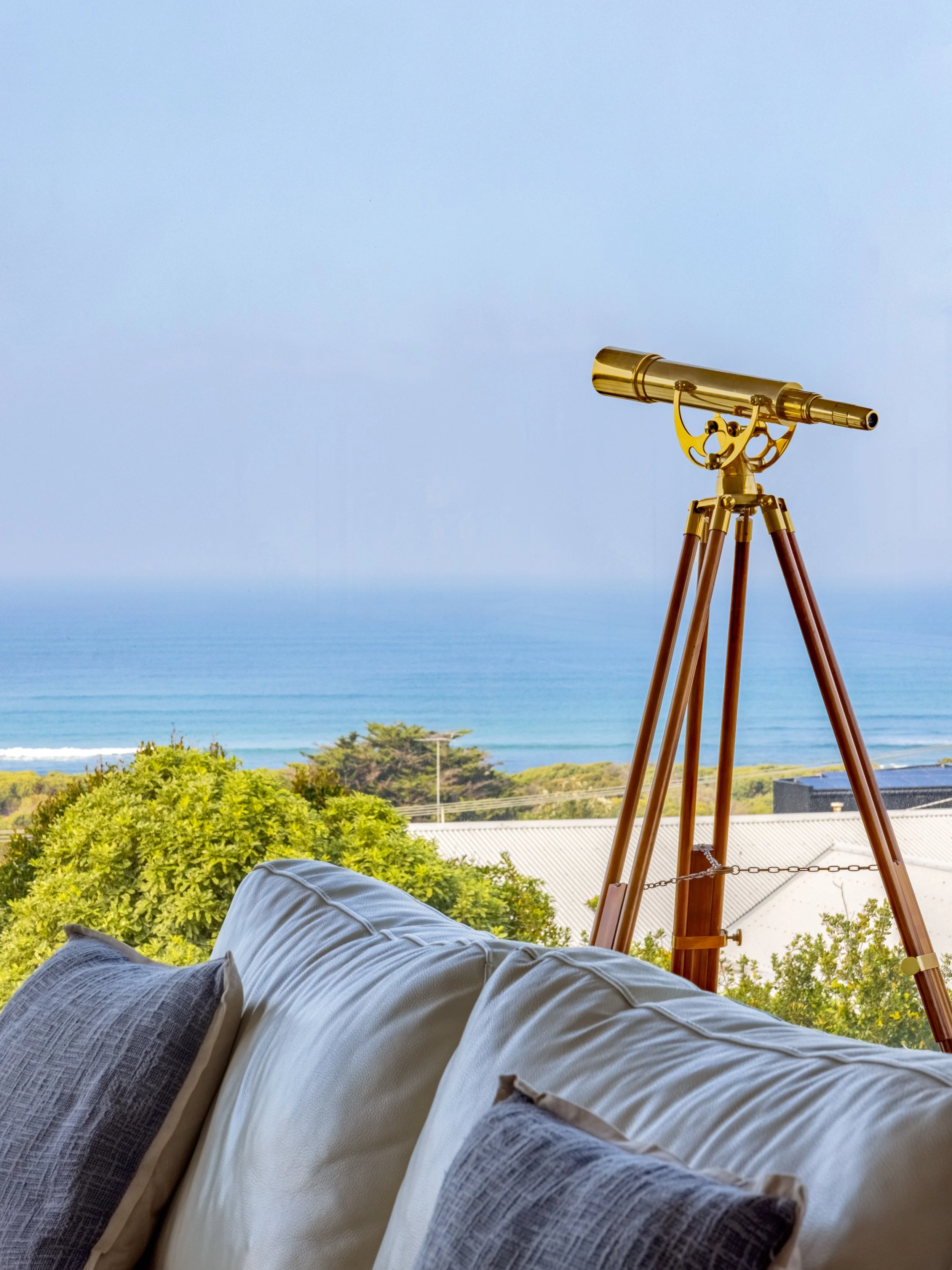 A person lying on a couch or bed, overlooking a scenic view of trees, a building with a metal roof, and a blue ocean with a clear sky. There is a golden telescope mounted on a wooden tripod directed towards the view.