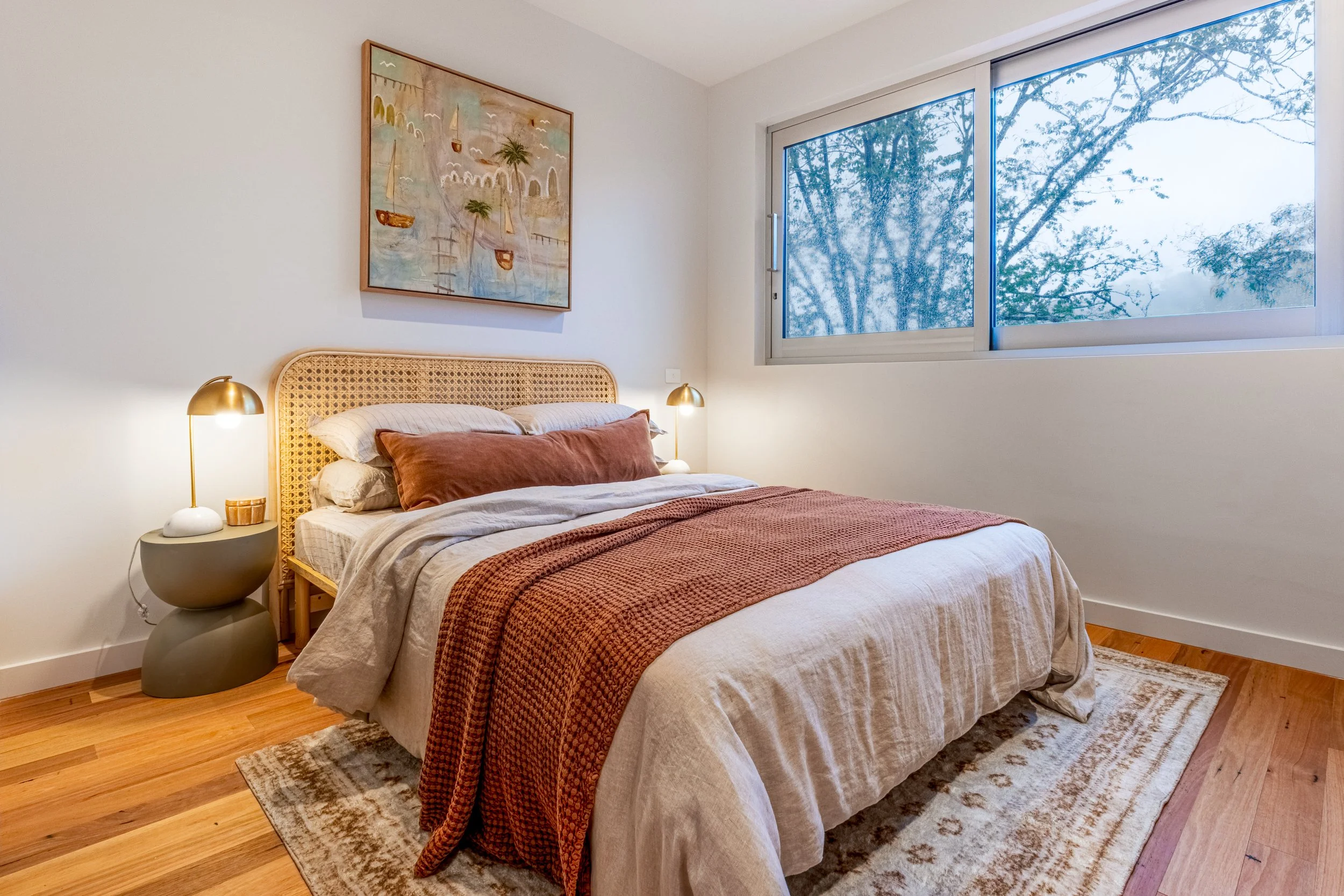 A cozy bedroom with a bed, two bedside tables with lamps, a painting above the bed, a large window with trees outside, and a patterned rug on wooden floor.
