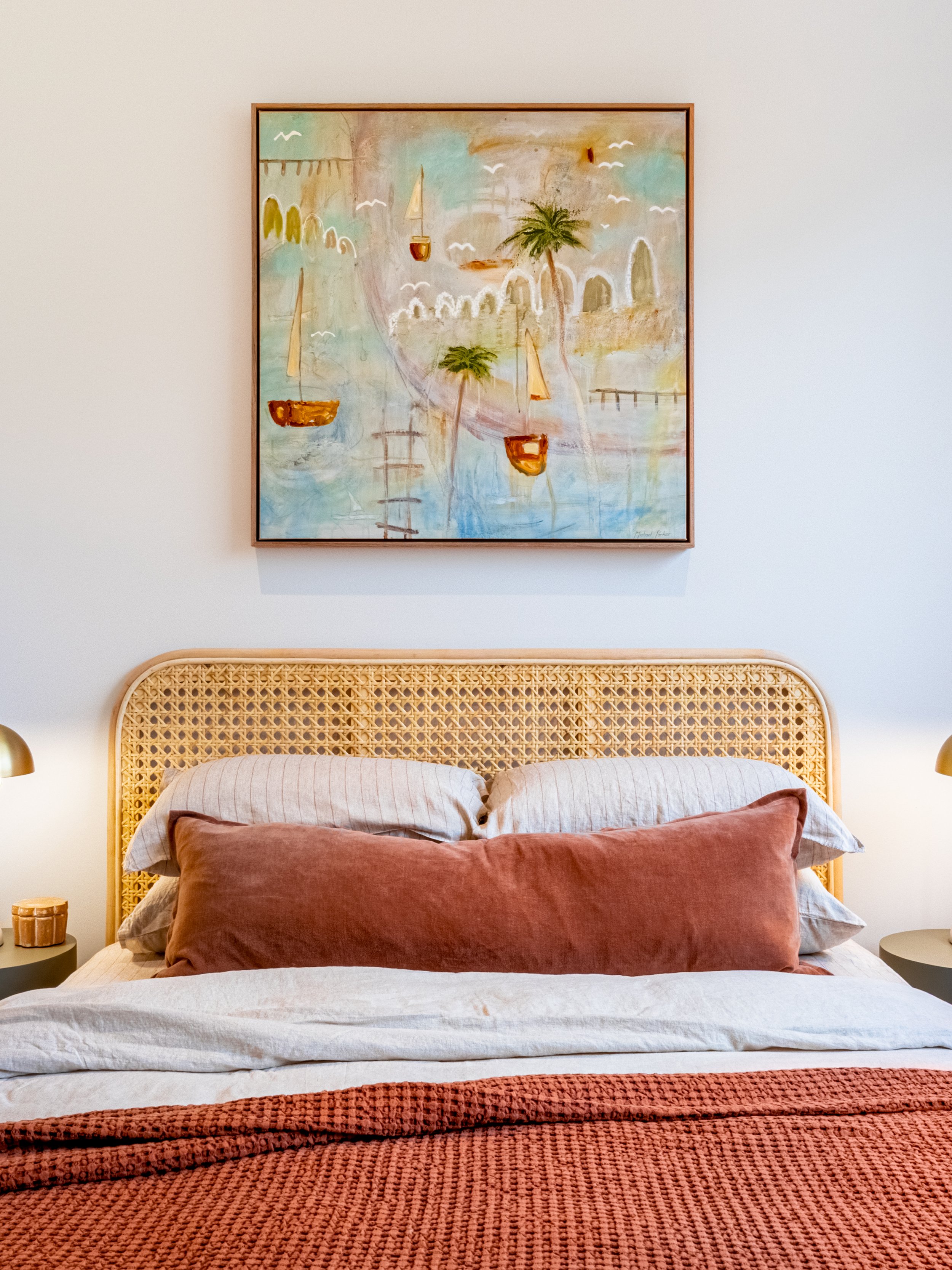 A bedroom with a bed featuring a natural woven rattan headboard, layered with beige and rust-colored pillows, a white bedsheet, and coral throw blanket at the foot. Above, a colorful abstract painting with sailboats, palm trees, and arched structures