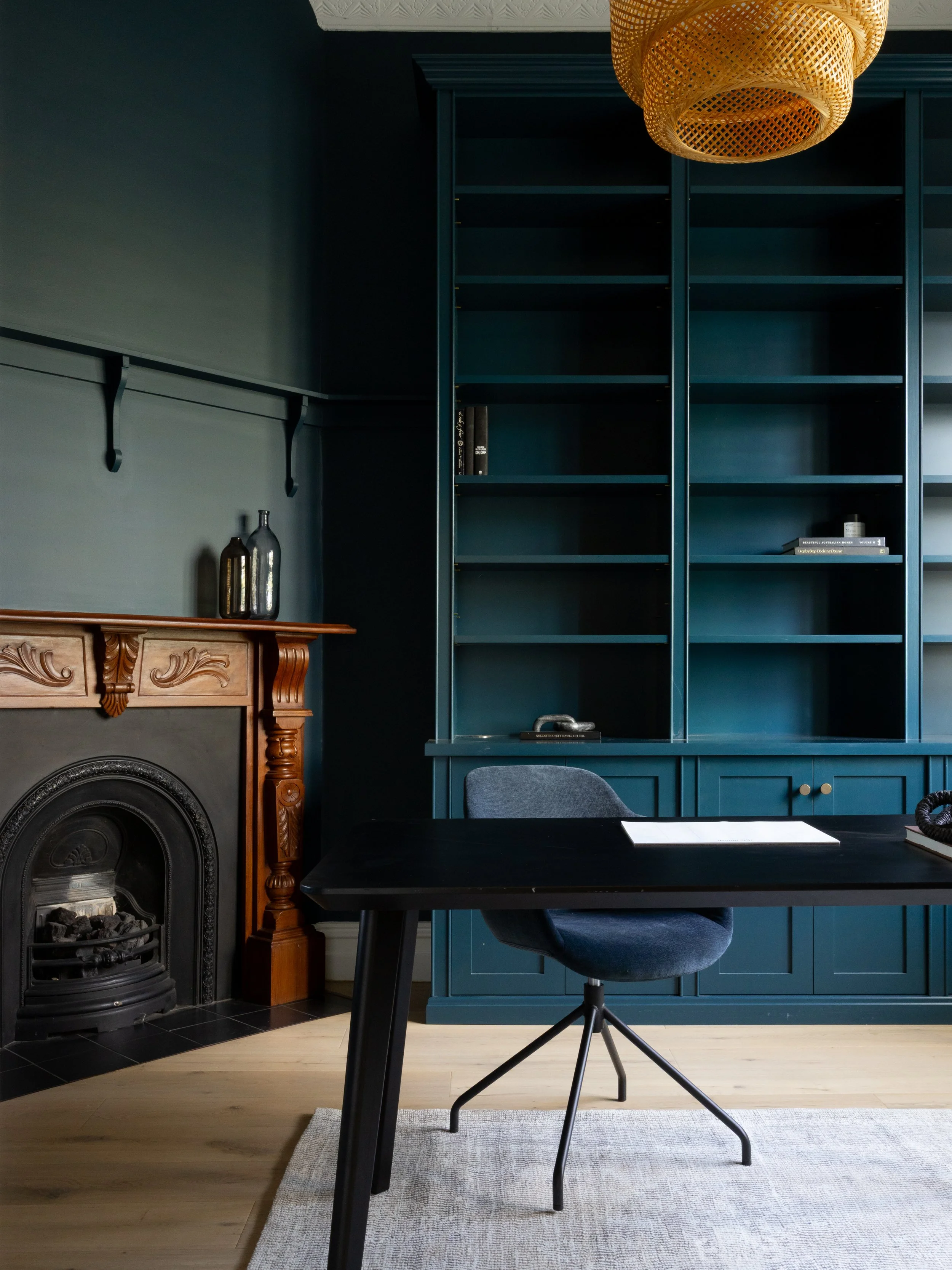 A room with a black table, a gray office chair, a blue bookshelf, a fireplace with wooden details, and decorative bottles on the mantel. There is a beige rug on a light wooden floor.