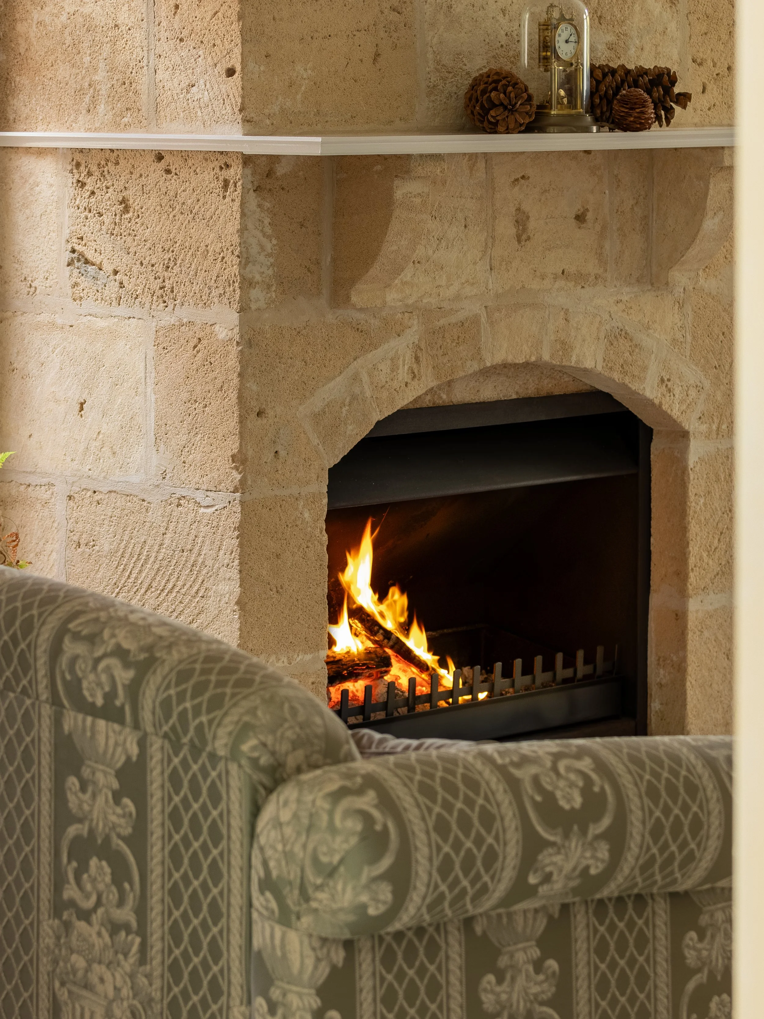 A fireplace with a stone surround, a fire burning inside, and a decorative armchair with patterned upholstery in the foreground. A shelf above the fireplace holds decorative pinecones and a vintage-style clock.