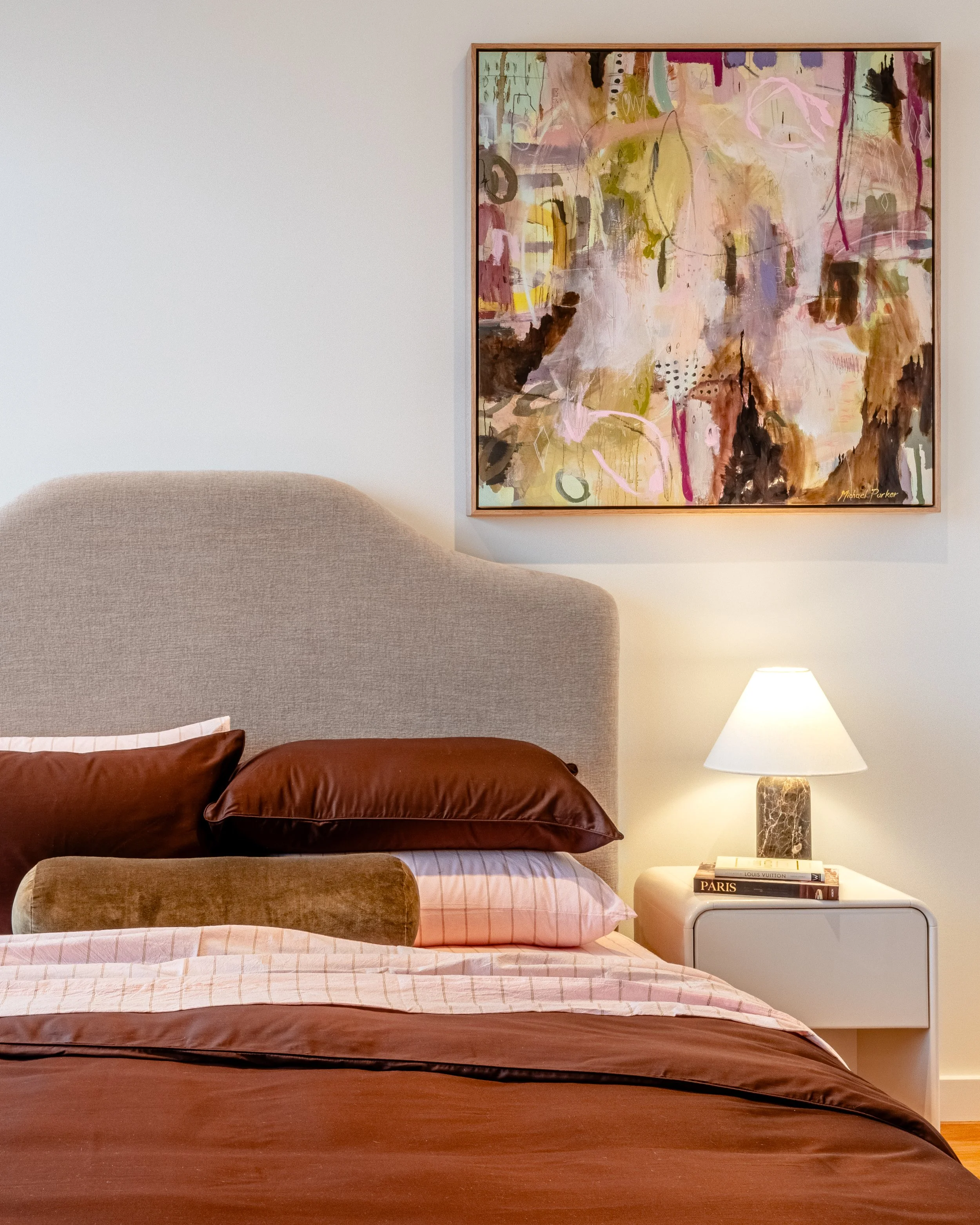 A bedroom with a beige upholstered headboard, a brown comforter, pink and brown checkered bedsheets, and two brown pillows. A white nightstand with a lamp, some books including one titled 'Paris', and a framed abstract painting with various pastel an