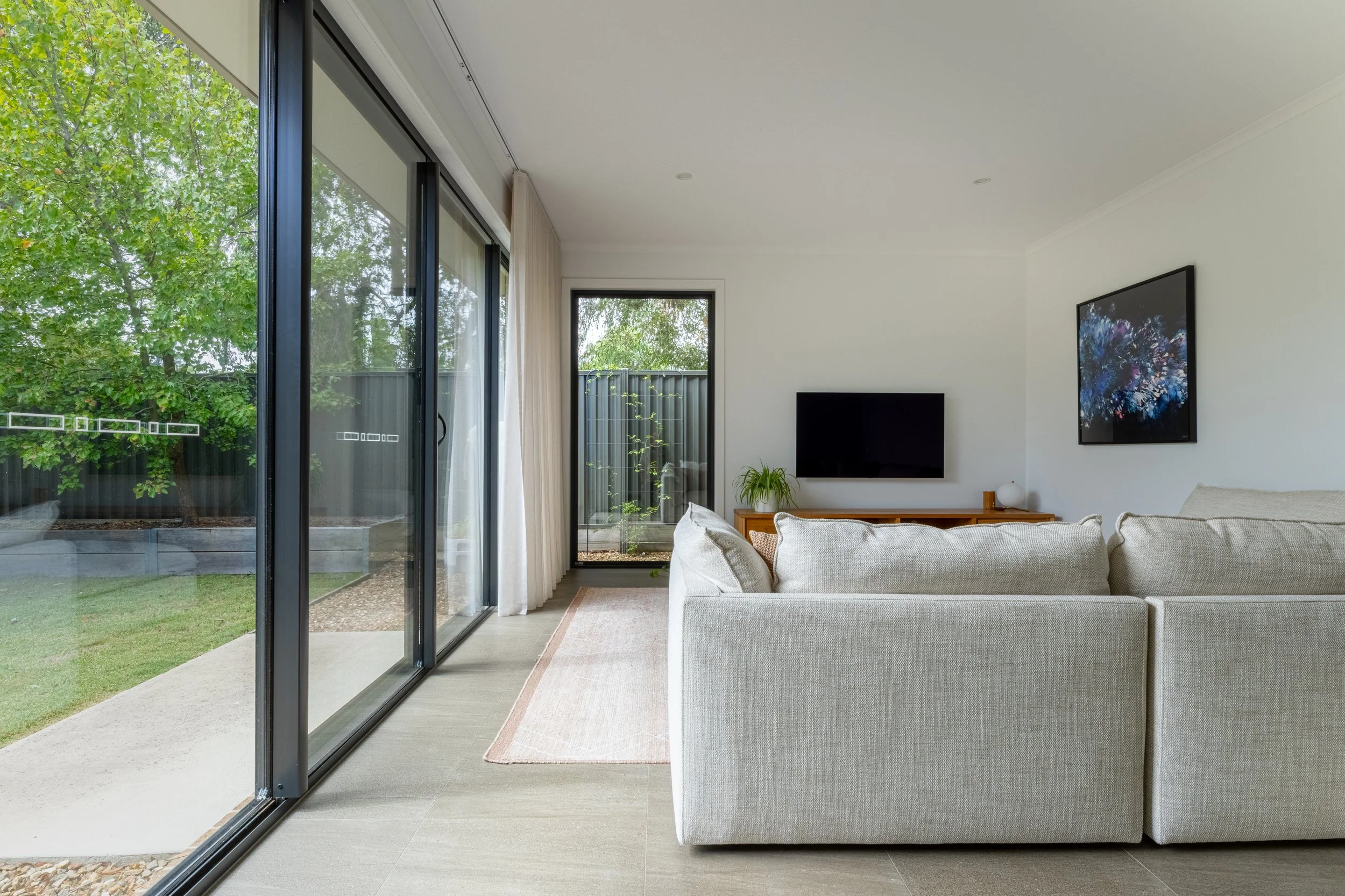 Modern living room with large glass sliding doors leading to a backyard with trees and a privacy fence. The room features a beige sofa, a wall-mounted flat-screen TV, a small wooden side table with a plant, and wall art, with a neutral color palette.