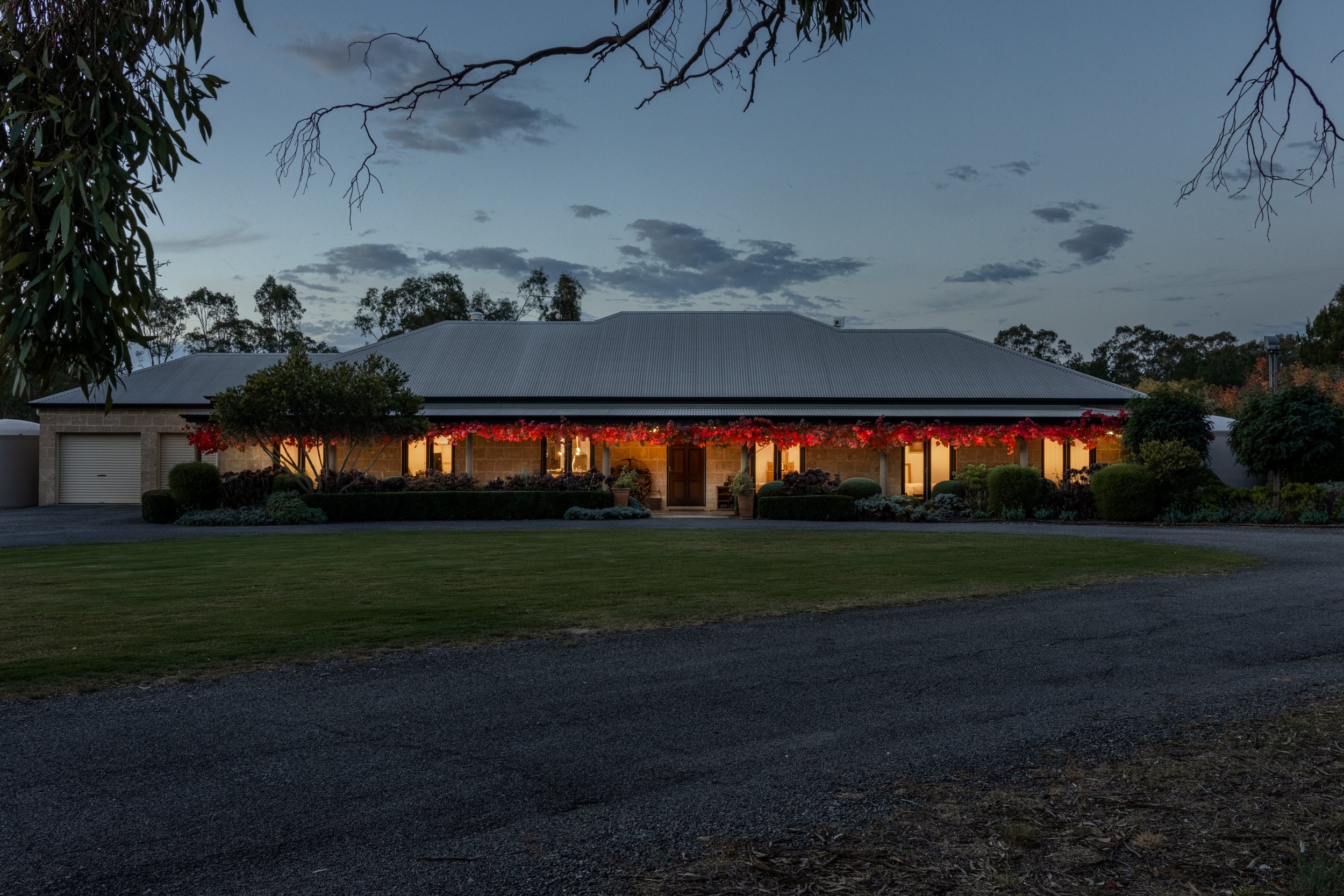 A house with a gray metal roof, brick walls, and a surrounding garden with trees and bushes, illuminated from inside with warm lights. The sky is dark with scattered clouds, and some branches are visible at the top of the image.