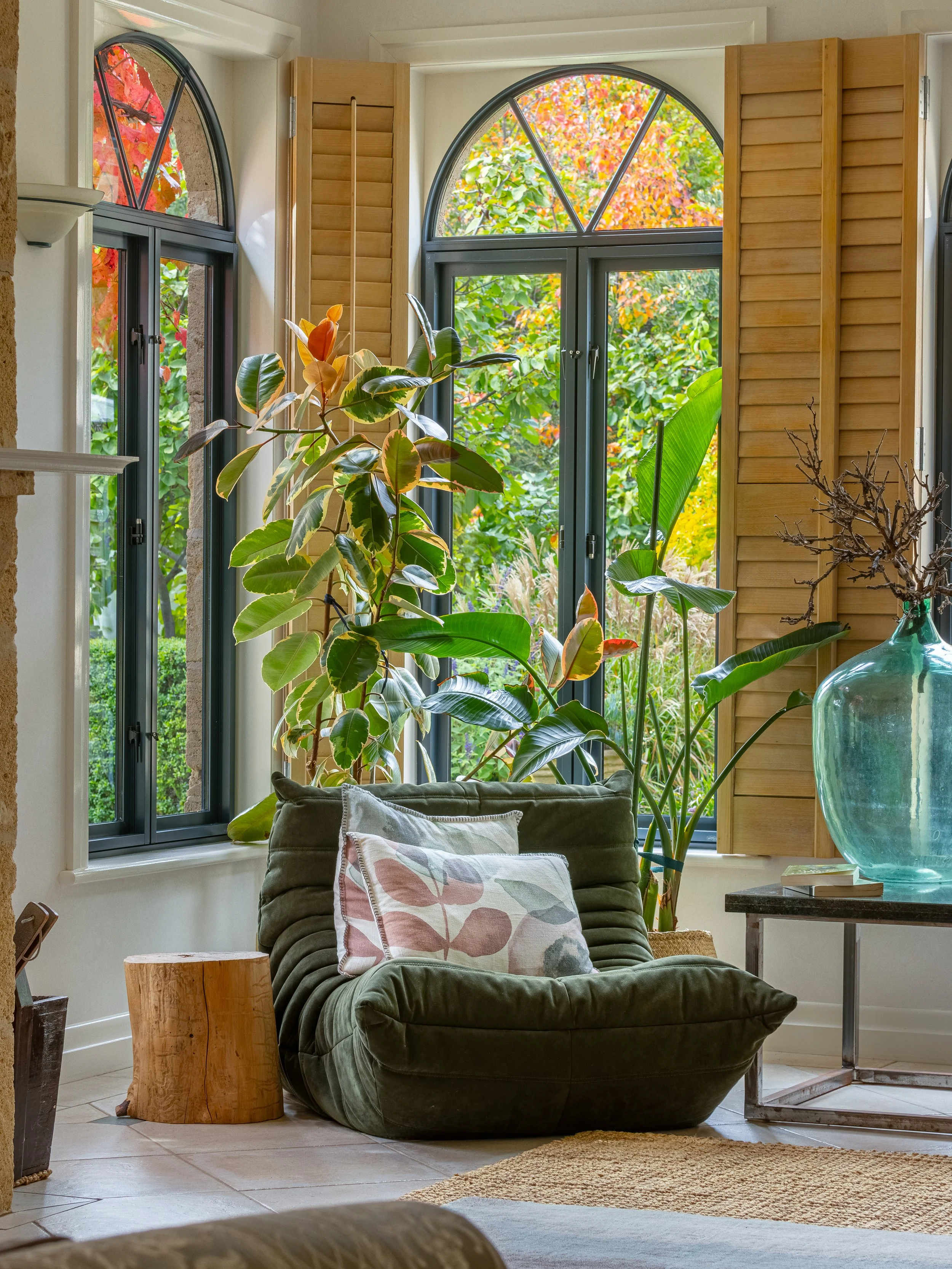 A cozy living room with large arched windows, wooden shutters, a green velvet armchair with a patterned pillow, a small wooden side table, tall plants near the window, and a large glass vase on a side table, with an outdoor garden view and colorful a
