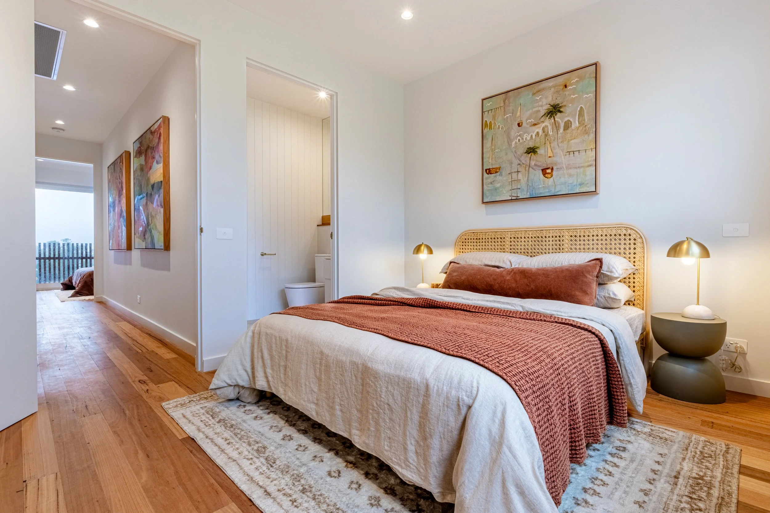 A bedroom with a bed featuring a rattan headboard, dressed in beige and rust-colored bedding. There are gold bedside lamps on either side of the bed. The wall above the bed has a large painting of a seascape with boats and palm trees. To the left, th