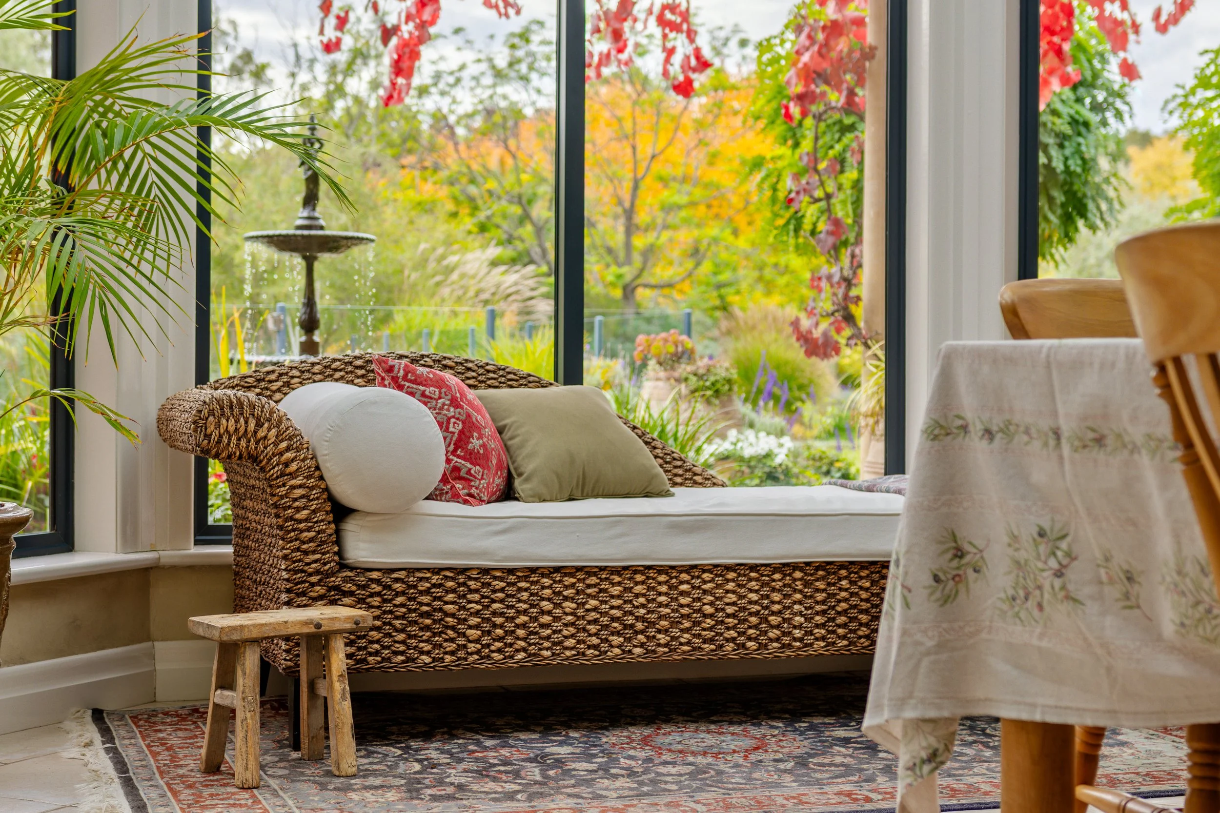 Living room with a wicker daybed, red and green pillows, and a small wooden stool in front. Large window displaying colorful fall foliage outside, with plants and a birdbath inside.