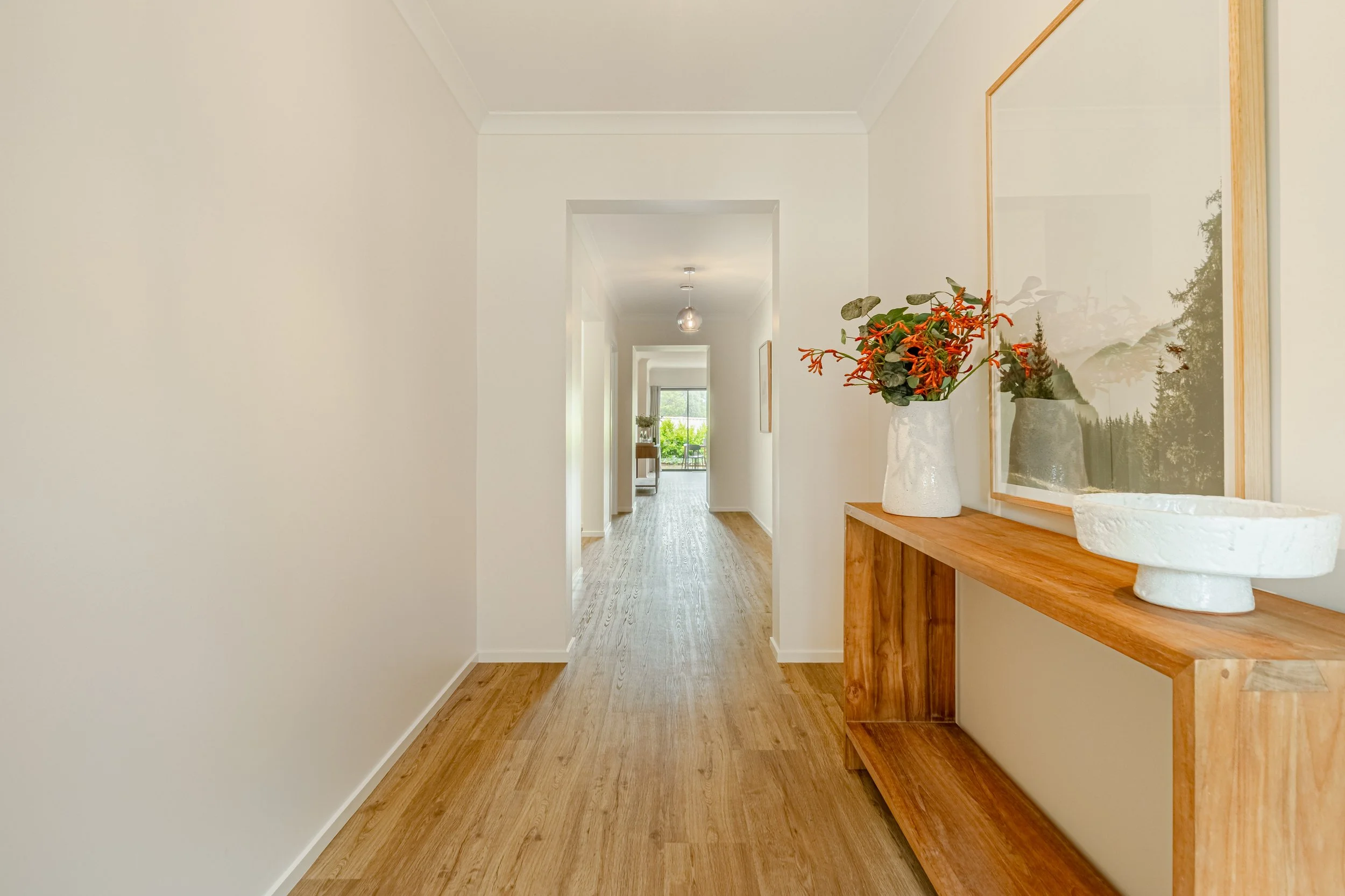 Interior hallway with wooden flooring, white walls, a wooden console table on the right with a large mirror and a vase with orange and green flowers. There's a window at the end of the hallway revealing greenery outside.