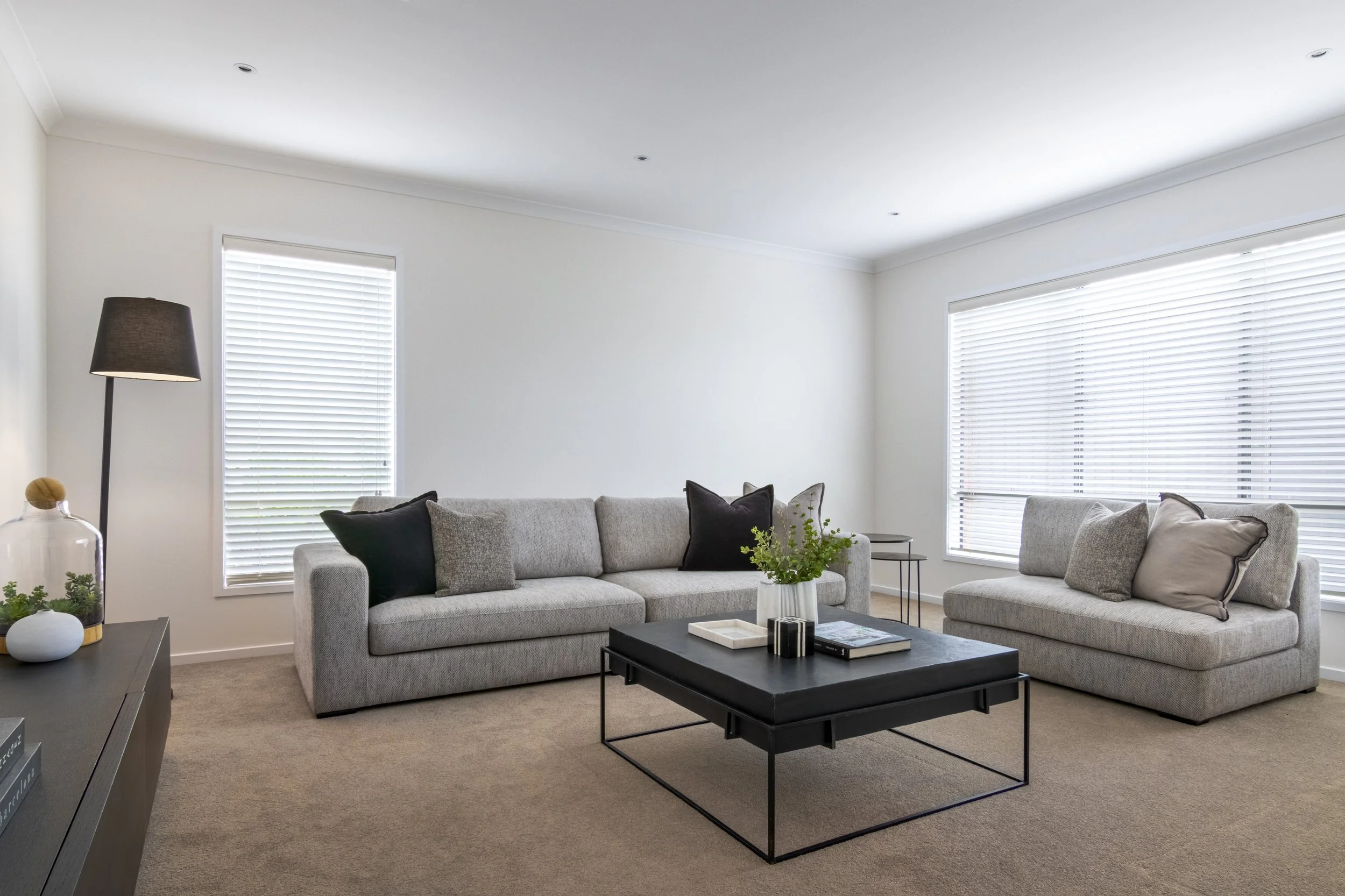 Living room with two gray sofas, black coffee table, side tables, black and beige pillows, large windows with blinds, and a black floor lamp.