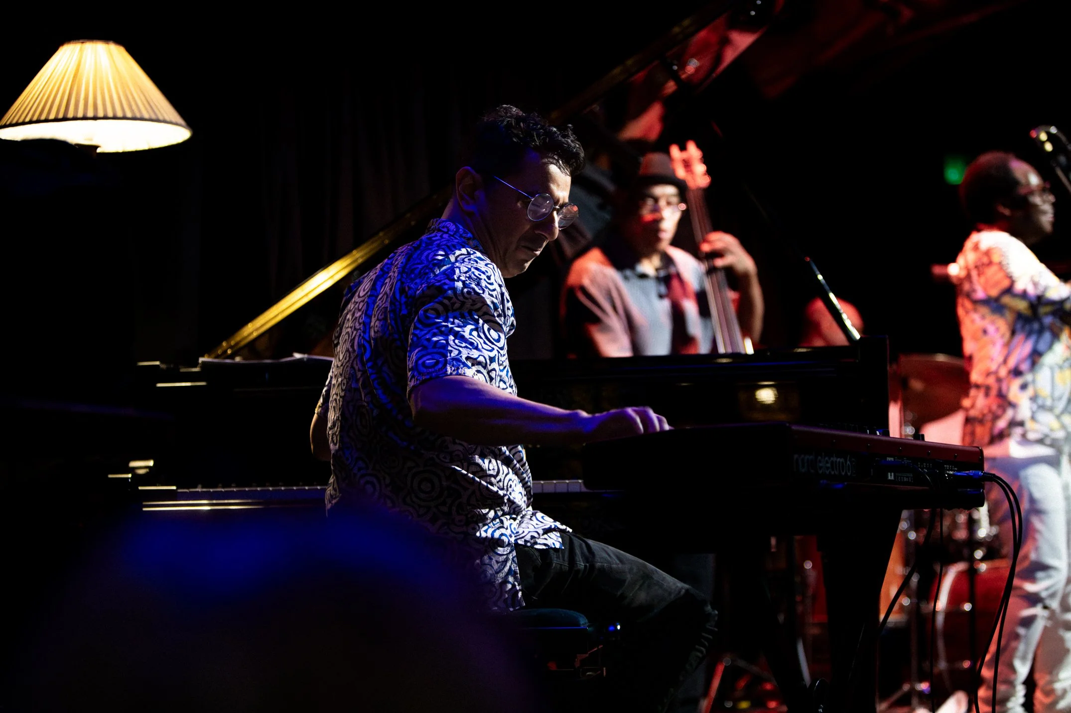A jazz band performing on stage with a pianist in the foreground, a double bassist in the background, and a vocalist to the right. The stage is dimly lit with warm lighting and a standing lamp.