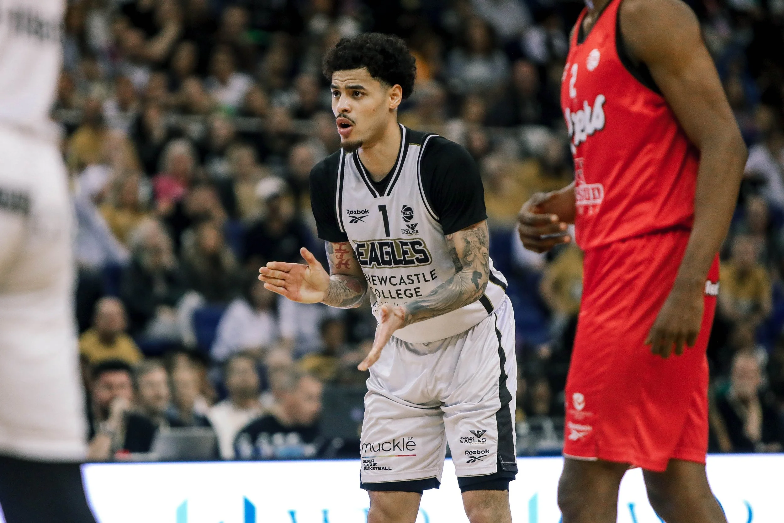 A basketball player wearing a black and white jersey with the text 'Newcastle College' and 'Eagles', standing on the court with a focused expression, clapping his hands, with another player in a red jersey partially visible beside him.