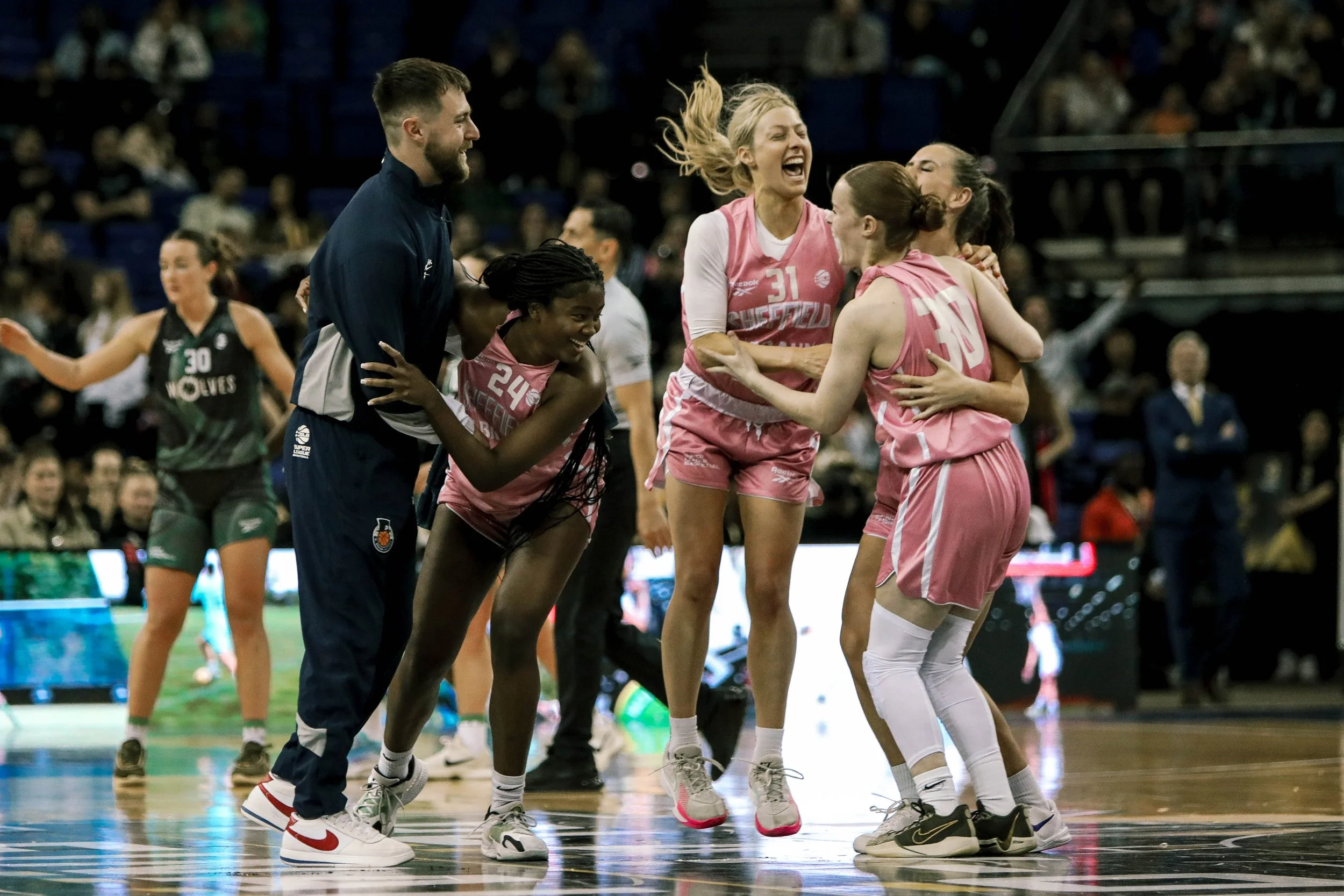 Group of female basketball players and a coach celebrating and hugging during a game on the court. Players in pink uniforms and one person in a dark uniform in the background.