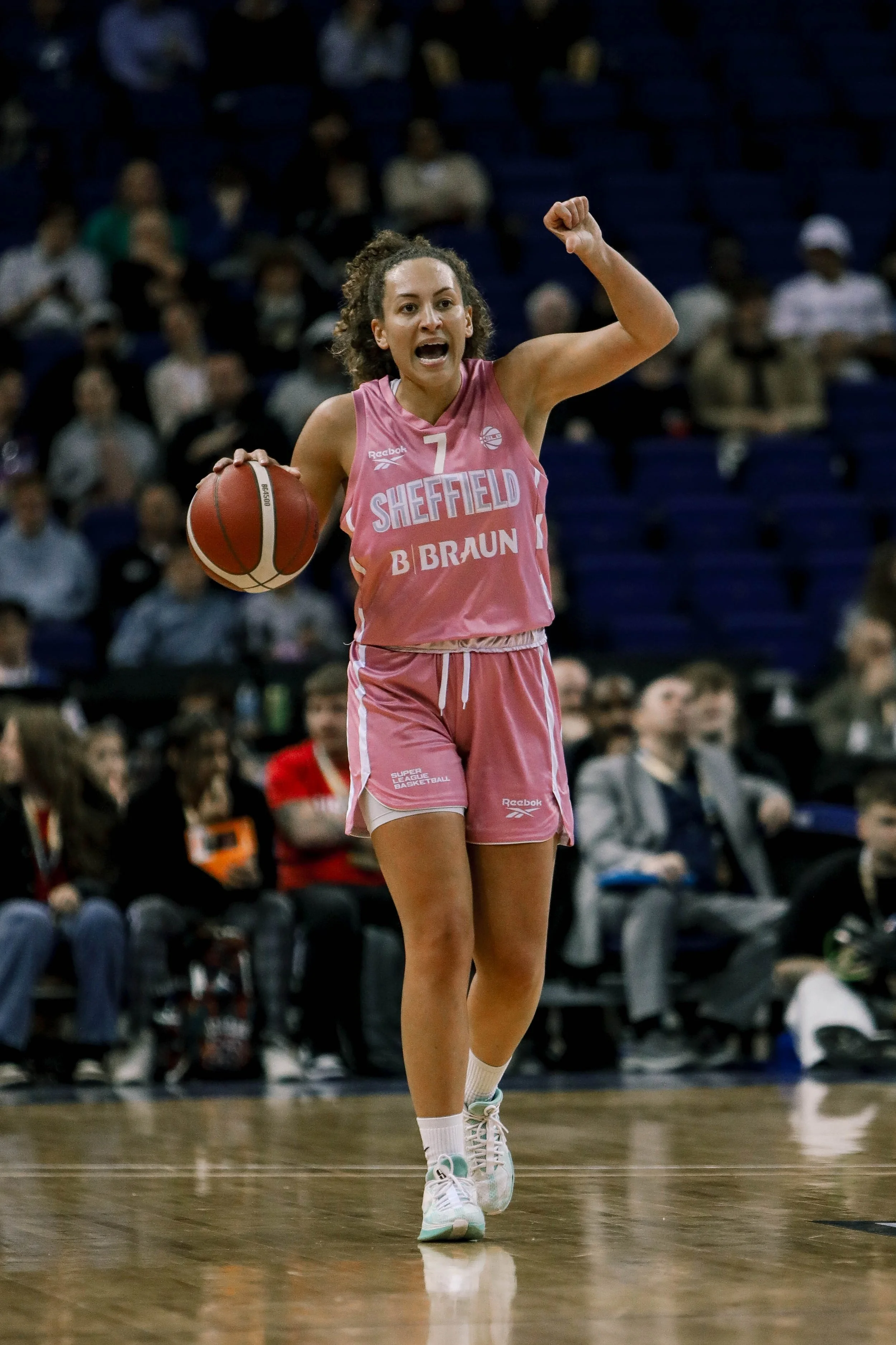 A female basketball player in a pink uniform with the number 7, holding a basketball, celebrating on the court during a game.