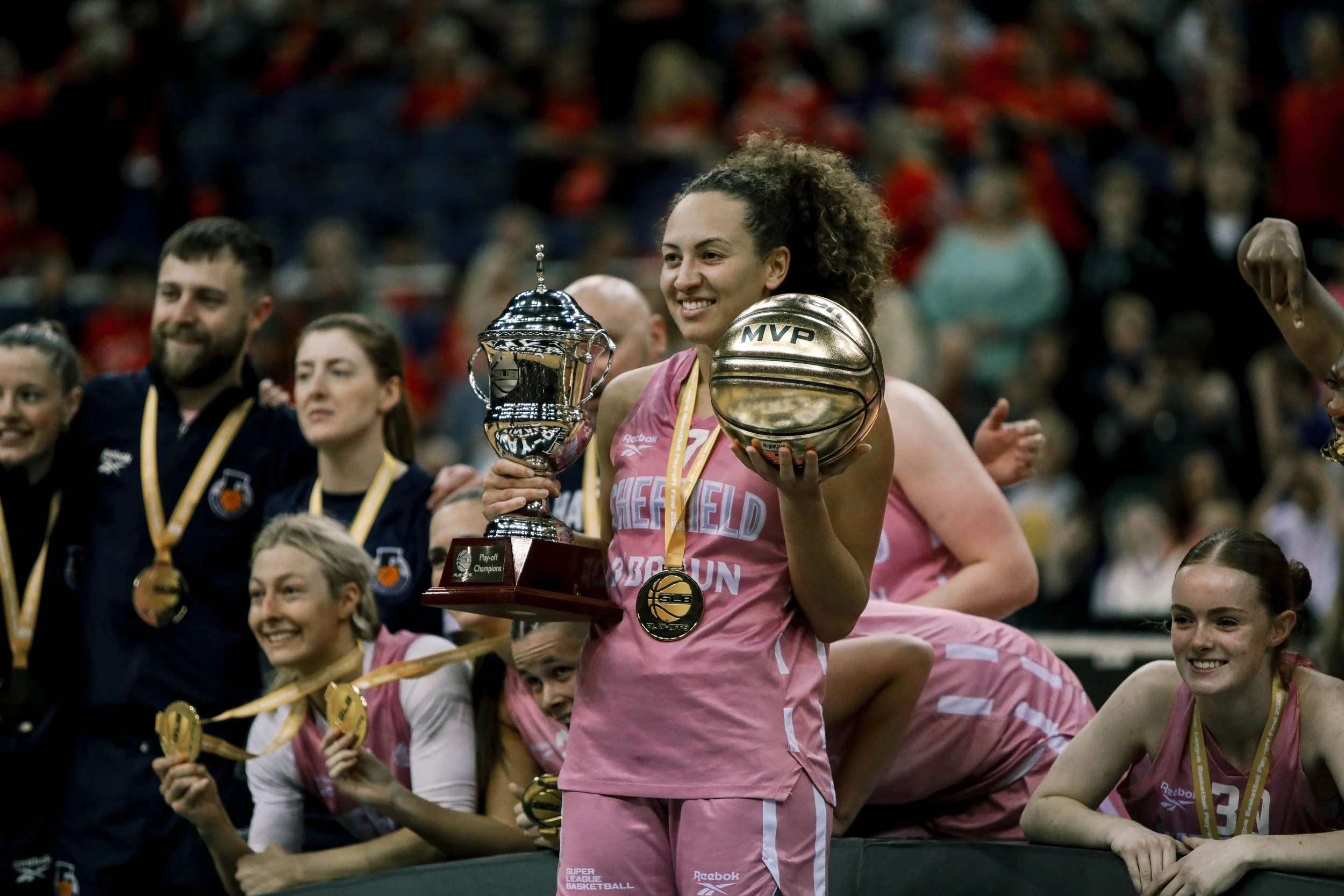 A female basketball player wearing a pink uniform holding a trophy and a gold basketball, surrounded by teammates and supporters celebrating.