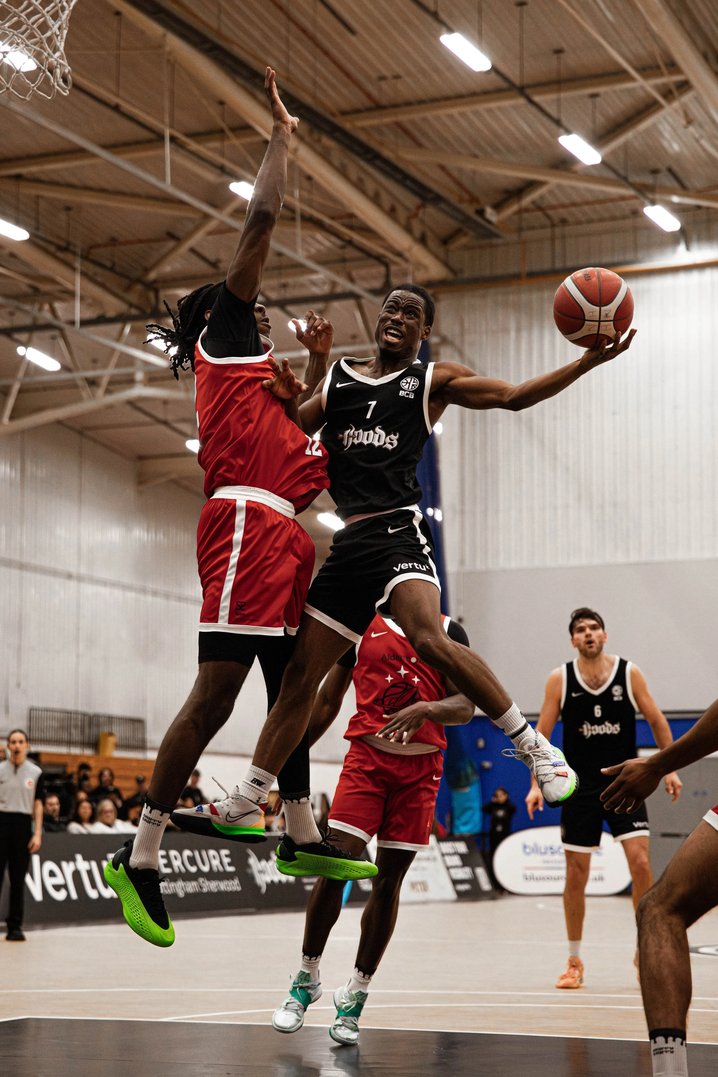 Two basketball players in black and red uniforms compete near the hoop, with one attempting to score while the other defends. The player in black is mid-air, reaching for the basket with the ball in hand. Other players and spectators are visible in t