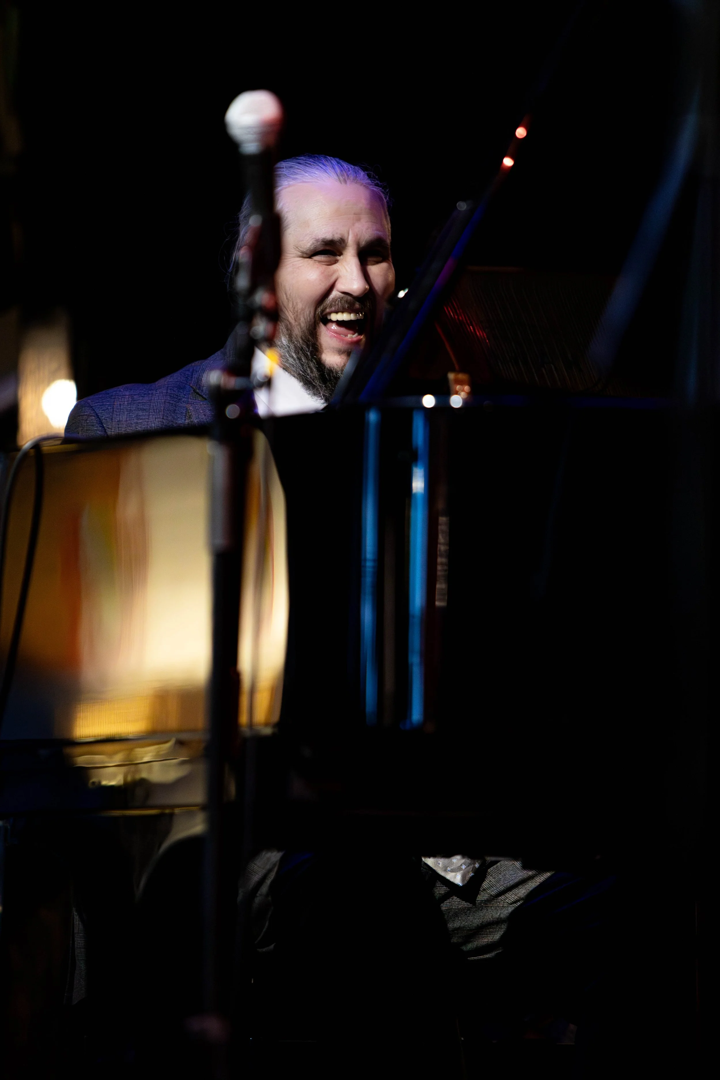 Man with long hair and beard smiling while playing the piano in a dimly lit setting during a performance.