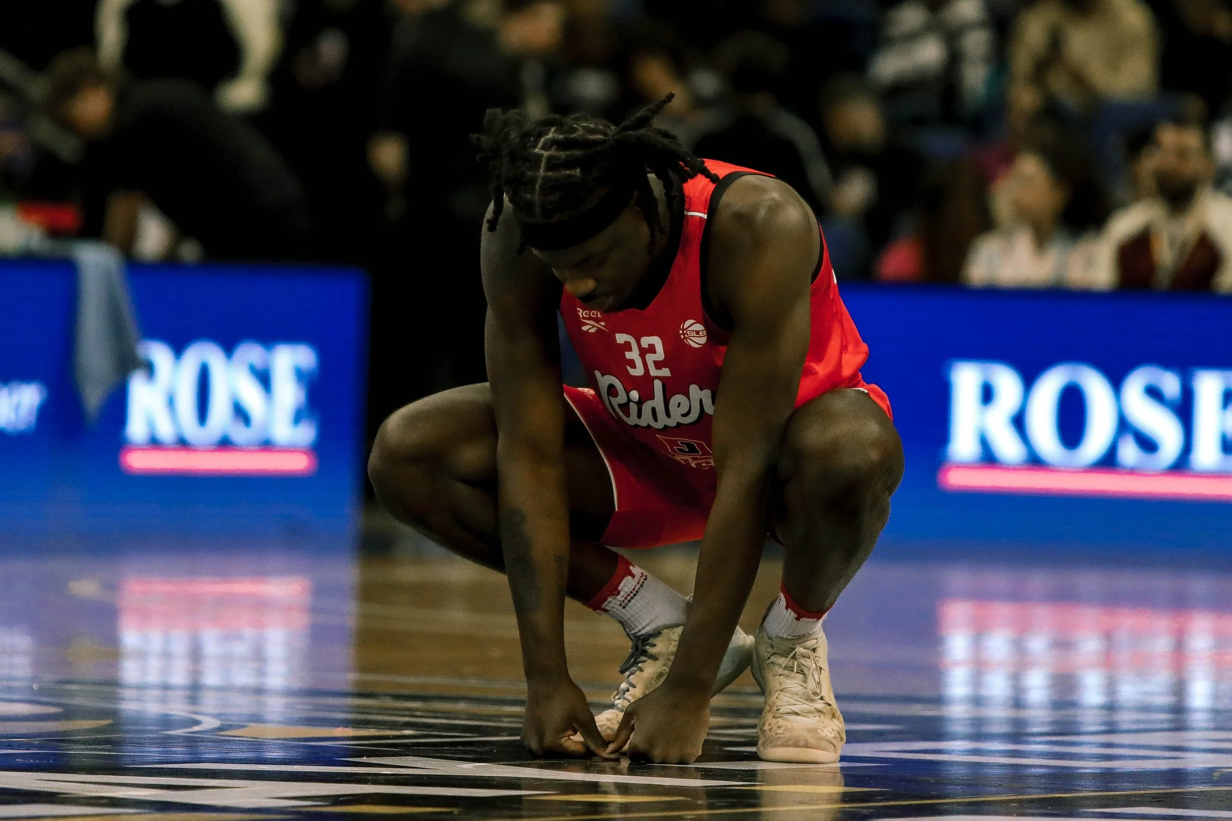 A basketball player in a red uniform with the number 32 is crouching on the court, adjusting his shoe. There are fans in the stands and digital advertising boards in the background.