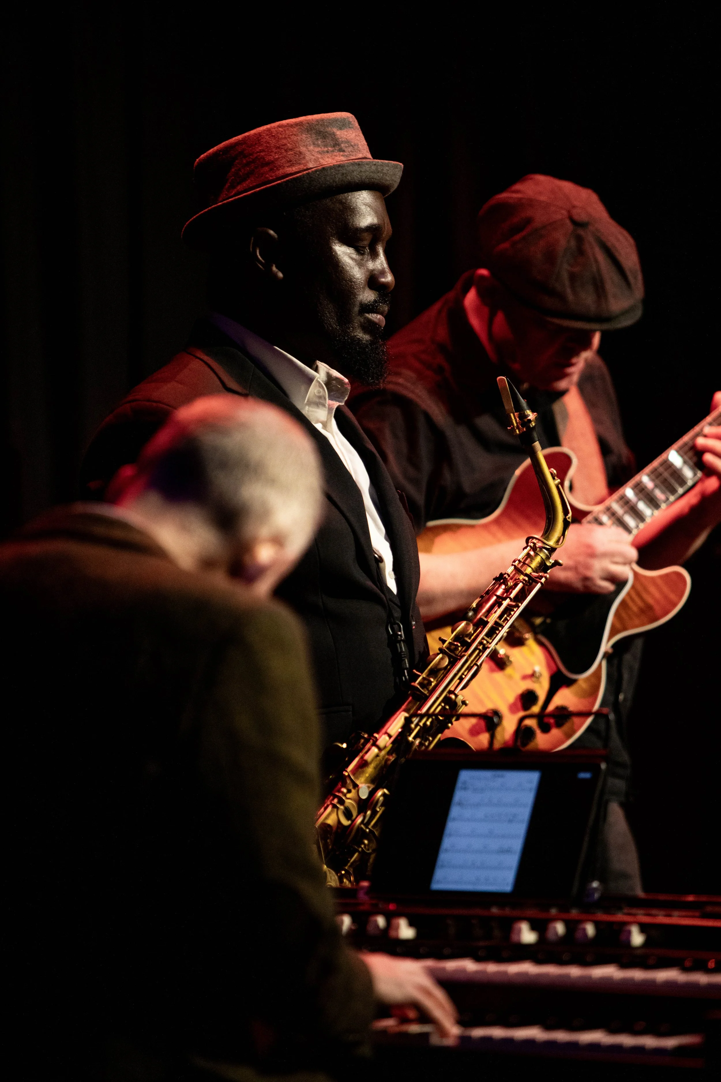 Jazz musicians performing on stage, including a man playing the saxophone and another man playing the guitar, with a keyboard in the foreground.
