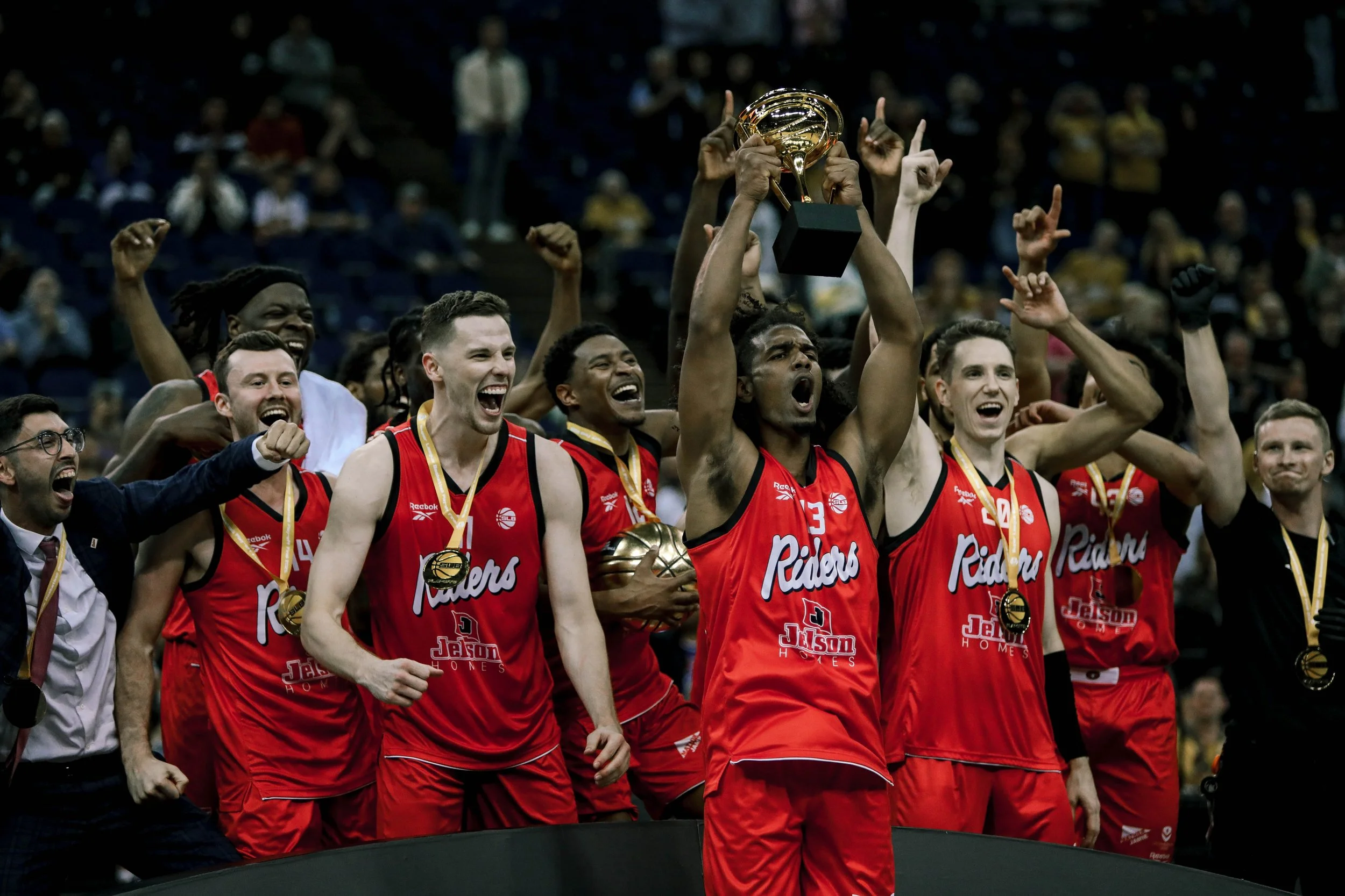 A basketball team wearing red jerseys celebrating with medals and holding a trophy after winning a game.