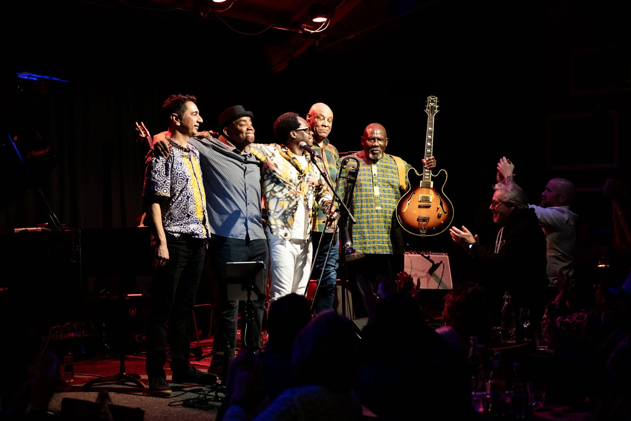 Jazz band performing on stage with five musicians, one holding a guitar, and two men clapping in front of the stage, in a dimly lit venue.