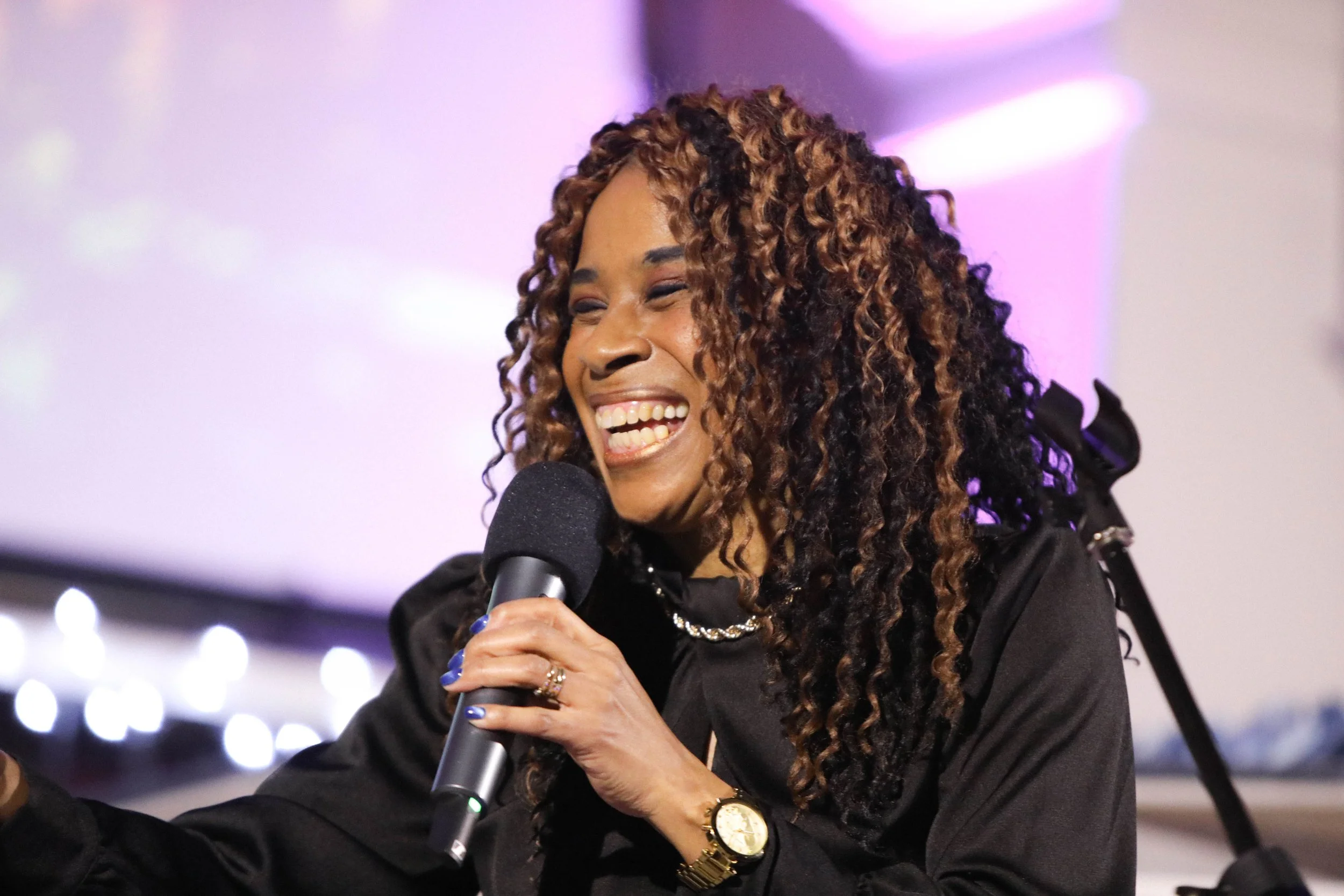 A woman with long, curly hair singing into a microphone and smiling, wearing a black outfit and a gold watch.