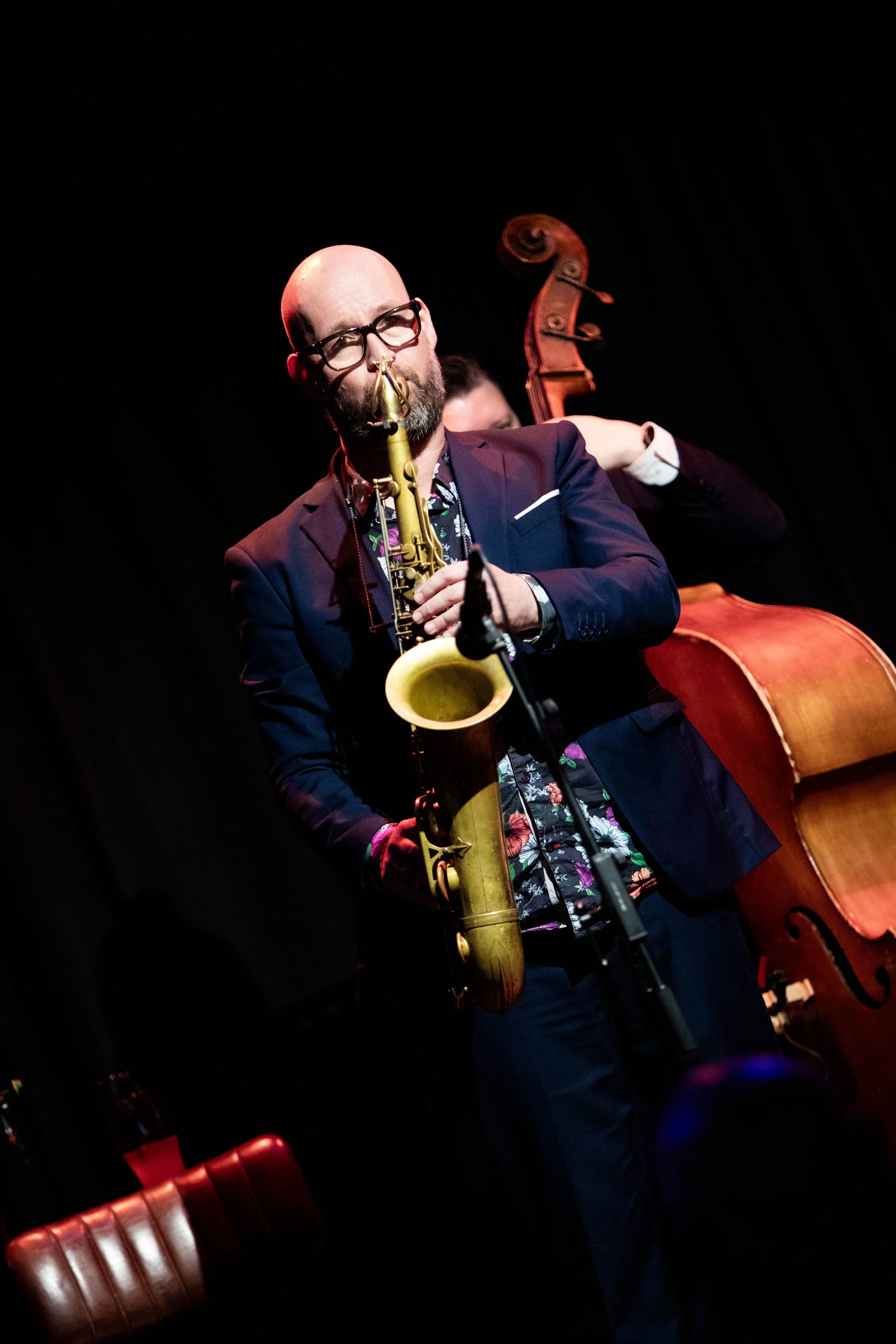 A man with glasses and a beard playing a saxophone on stage, with a double bass in the background.