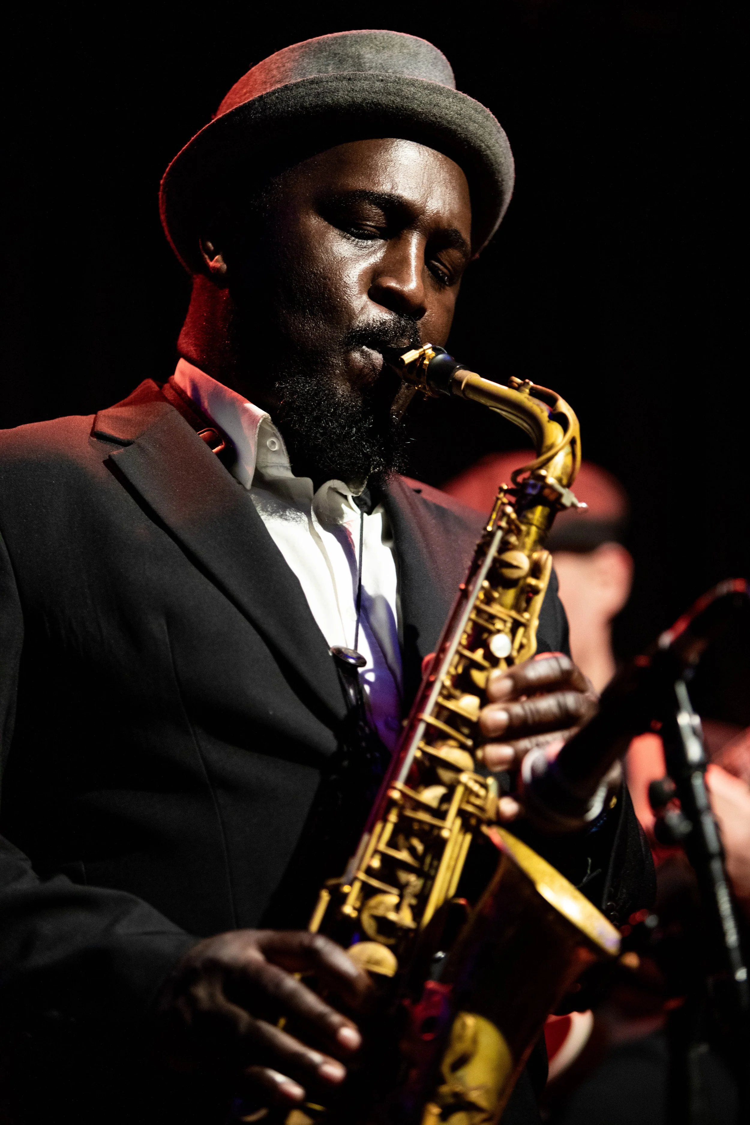 A man with a beard, wearing a gray hat and a black suit, playing a saxophone on stage during a performance.