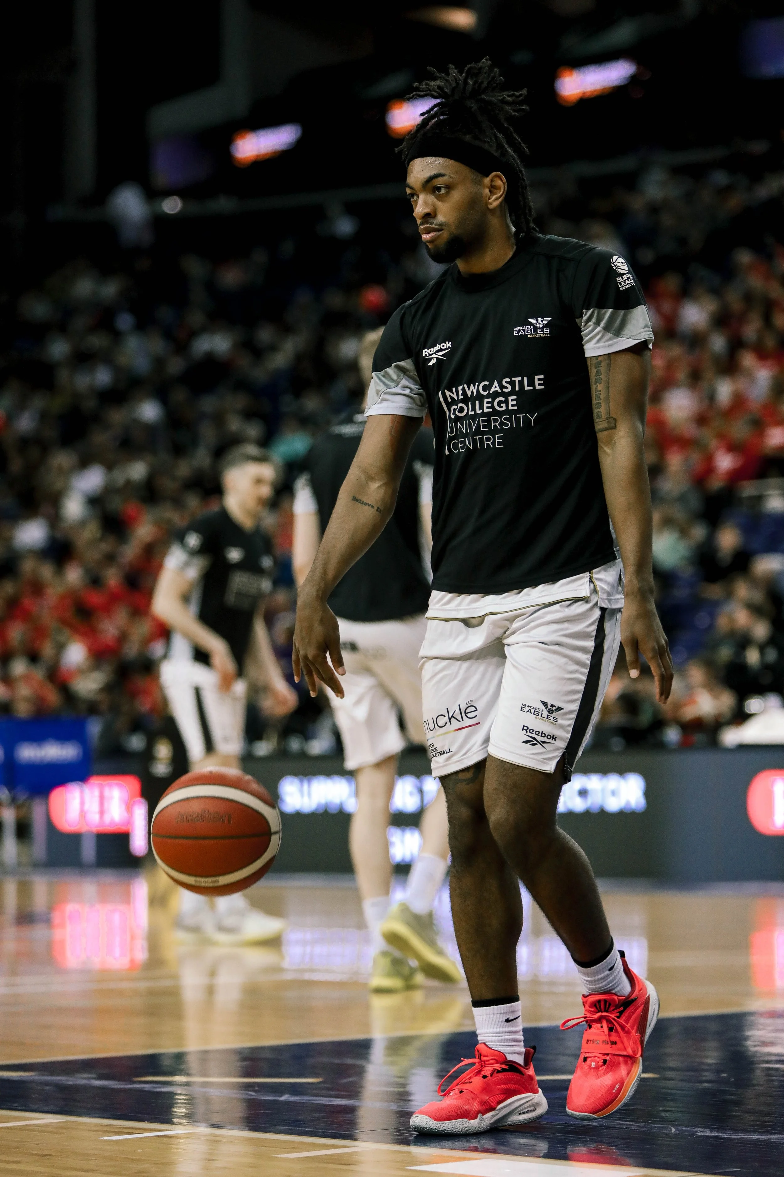 A basketball player in black and white uniform practicing on a court with a red basketball, wearing red sneakers, a black headband, and tattoos on his arms, during a game or practice.