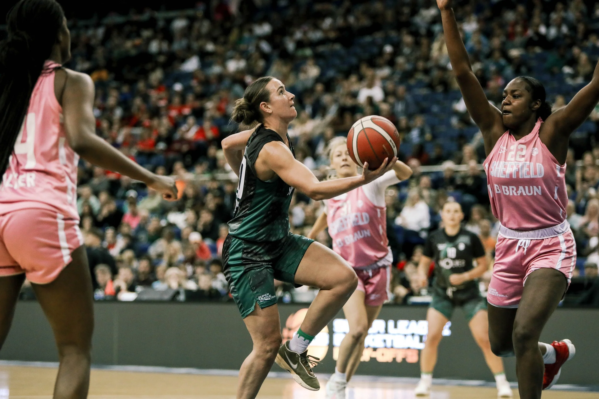Women playing basketball on indoor court, one in black uniform holding the ball, surrounded by opponents in pink uniforms, crowd in stands.