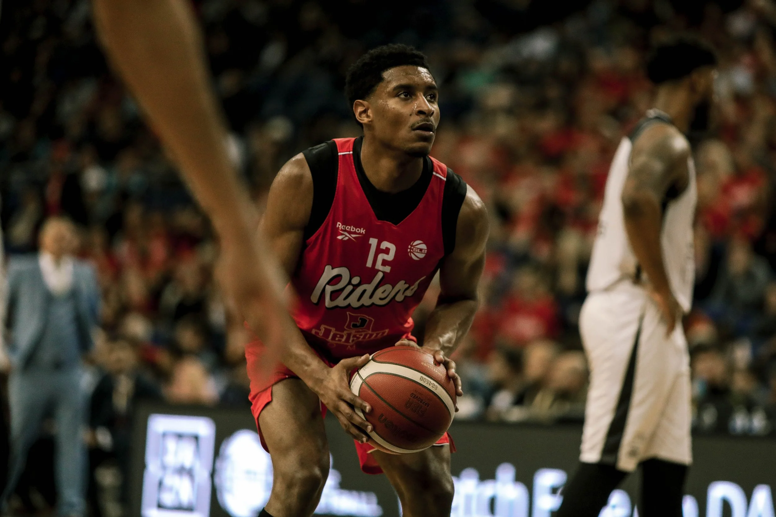 A basketball player in a red uniform with the number 12 is holding a basketball and preparing to shoot on the court. Another player in a white uniform is visible in the background, and there is a blurred crowd watching the game.