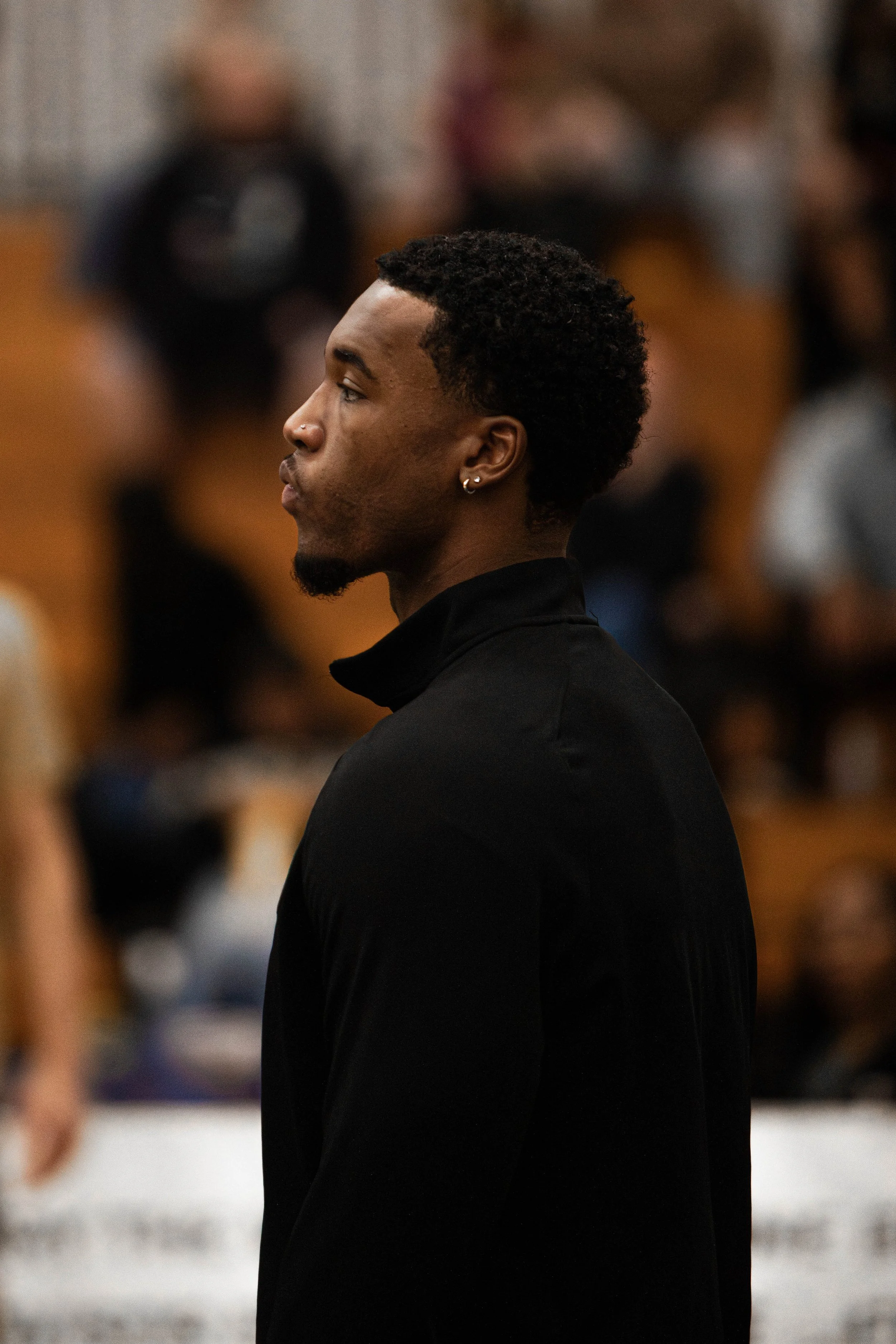 A young man with dark, curly hair, earrings, and a small goatee, wearing a black jacket, stands in profile indoors with a blurred crowd background.