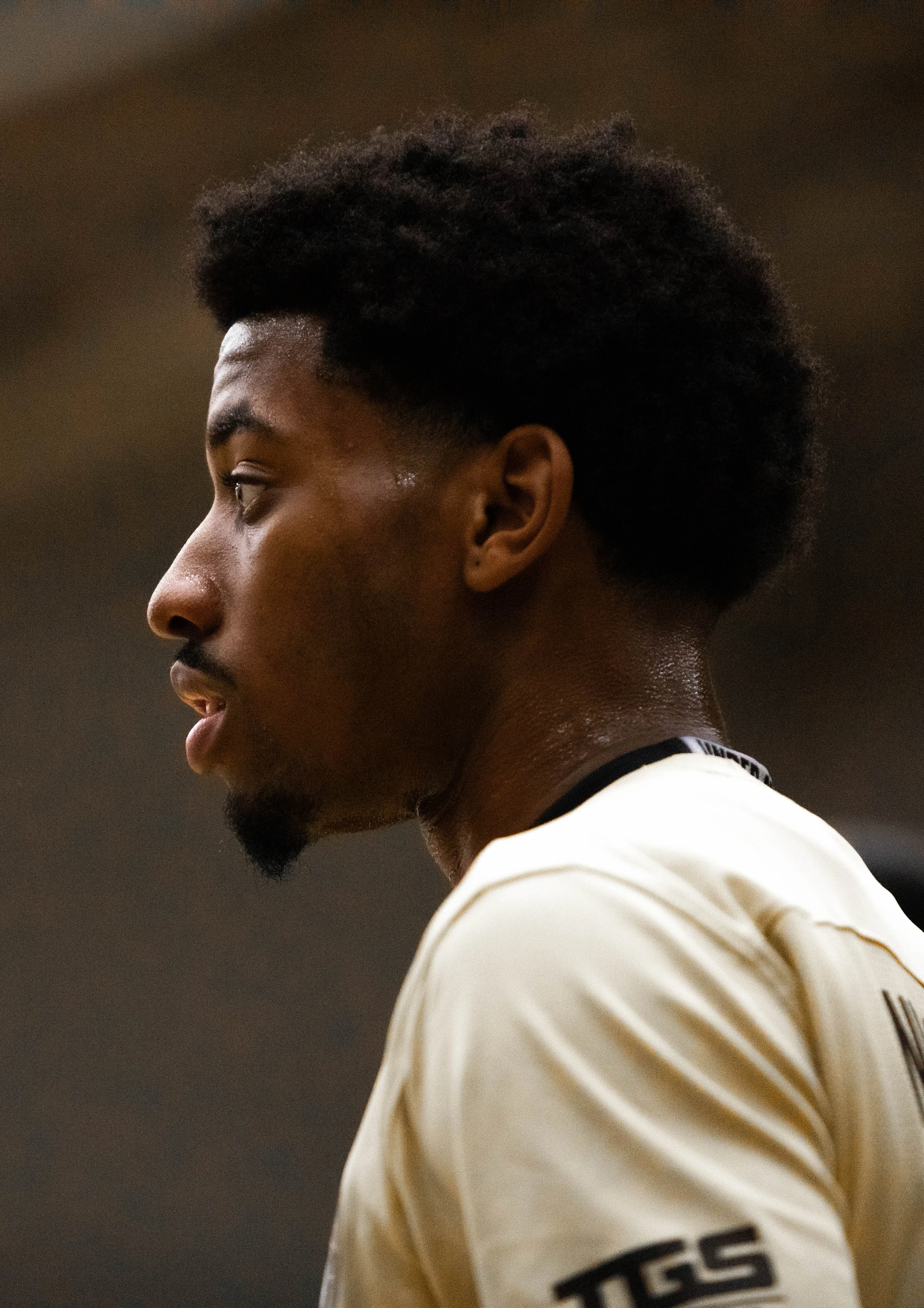 Close-up side profile of a young man with dark skin and black hair, wearing a cream-colored sports jersey with the word 'TESS' on the sleeve, looking serious or focused.