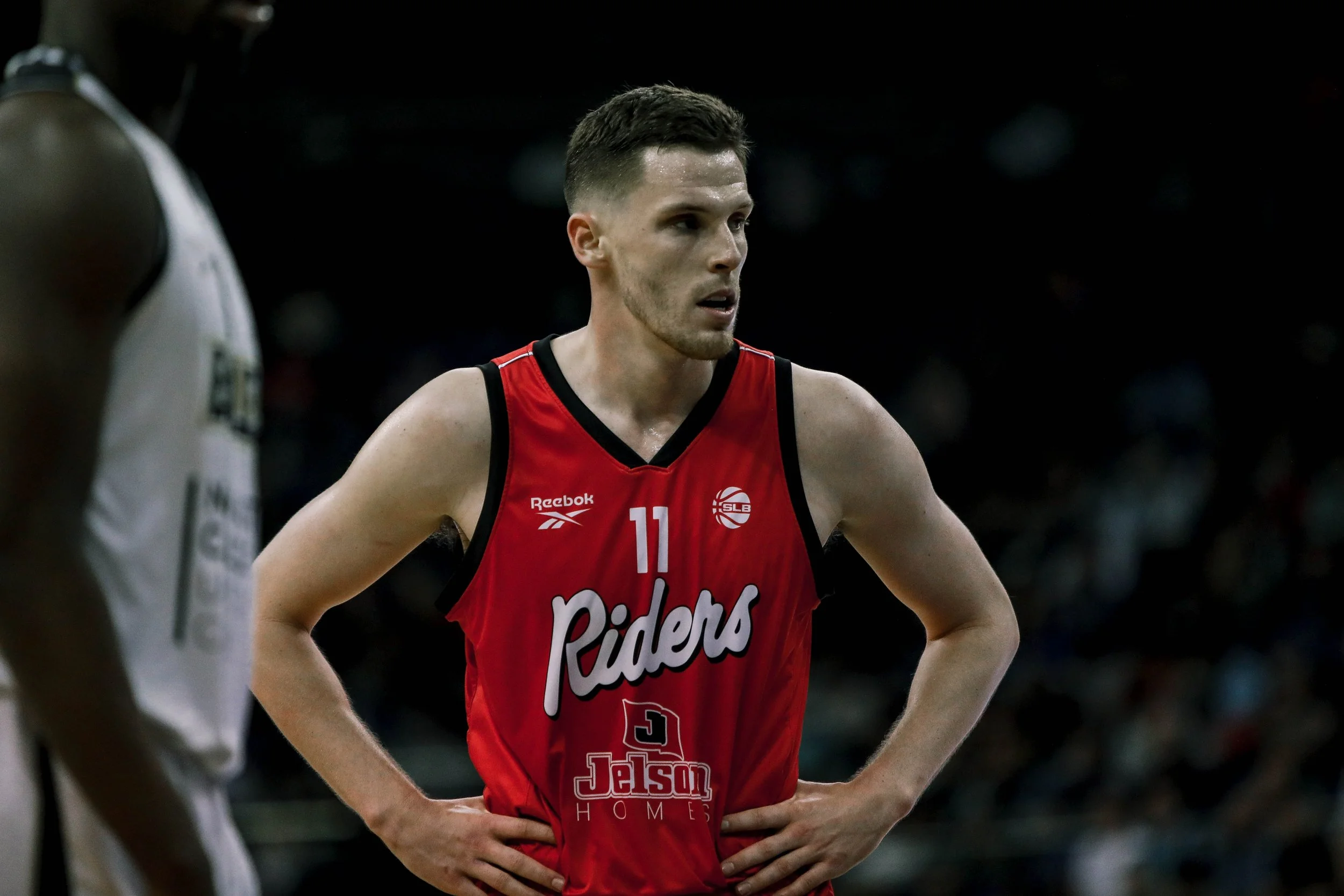 A basketball player wearing a red jersey with the number 11 and the word 'Riders' stands on the court with his hands on his hips, looking focused.