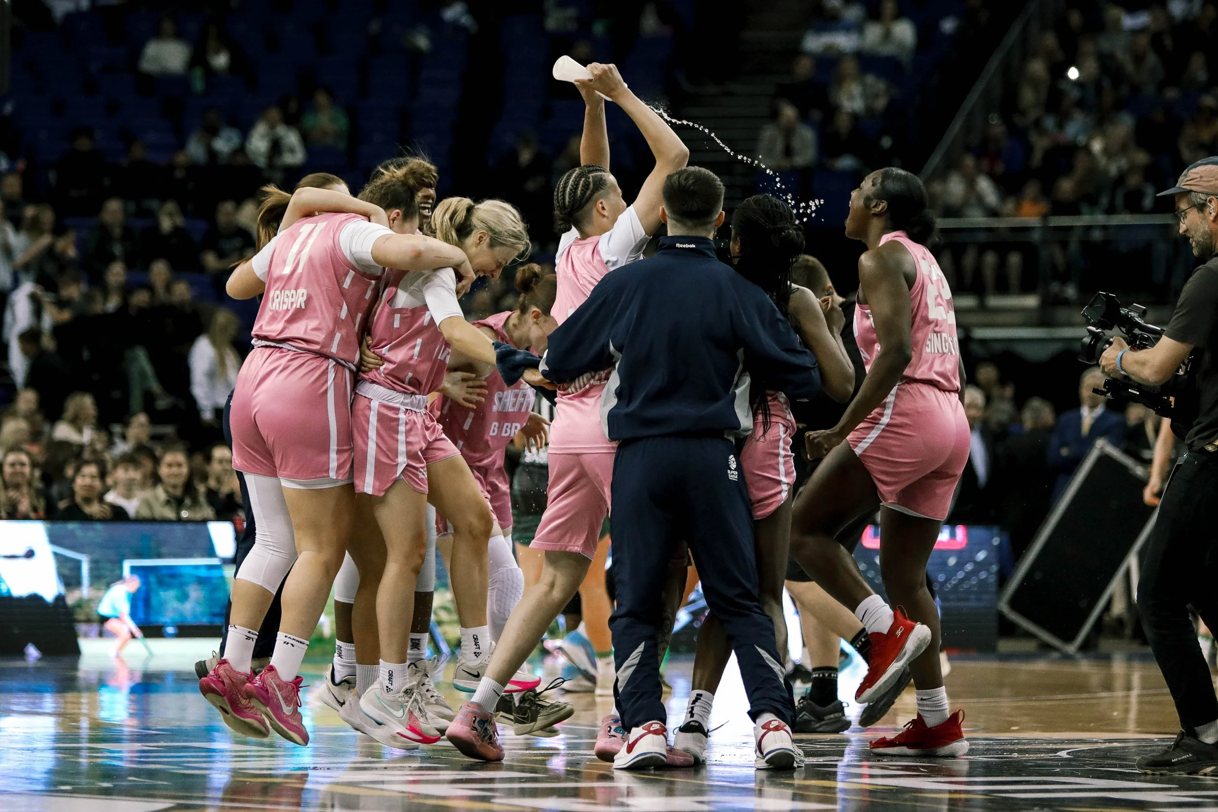 Women's basketball team celebrating on the court, some players are jumping and others are gathering around their coach, with a crowd watching in the background.