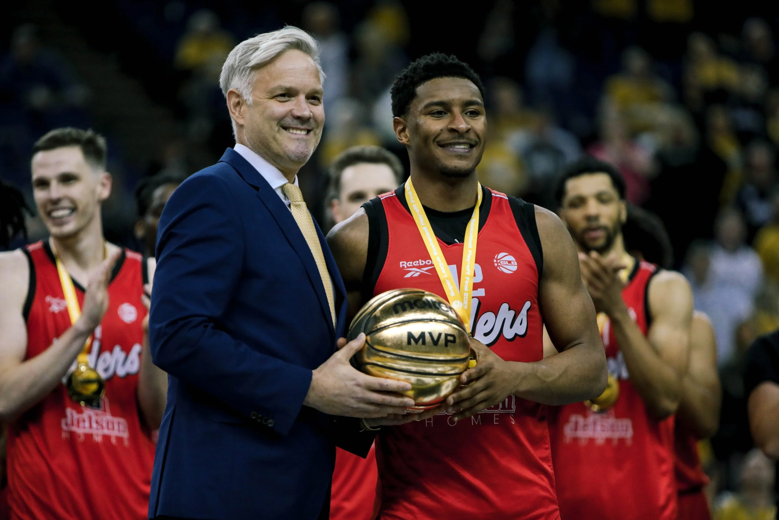 A man in a blue suit presents a gold MVP basketball to a basketball player in a red uniform during a celebration. The player is smiling and holding the basketball, with teammates and spectators in the background.
