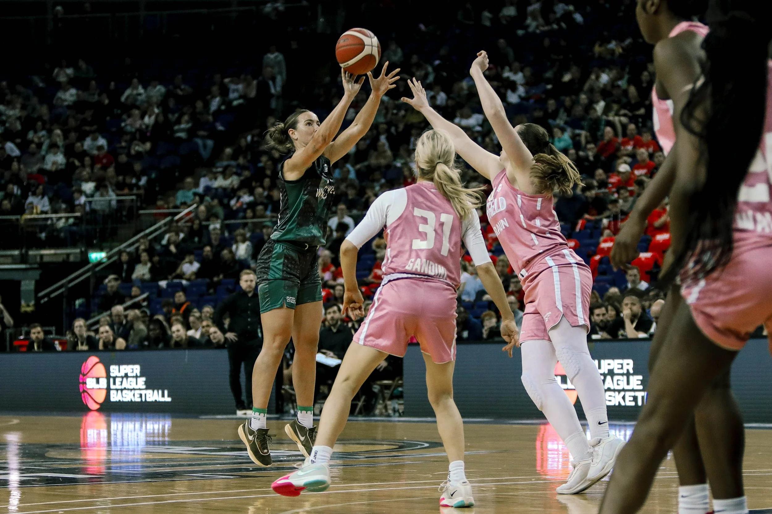 Women playing basketball in a stadium, one player in black jumping to shoot or pass the ball while other players in pink attempt to block.