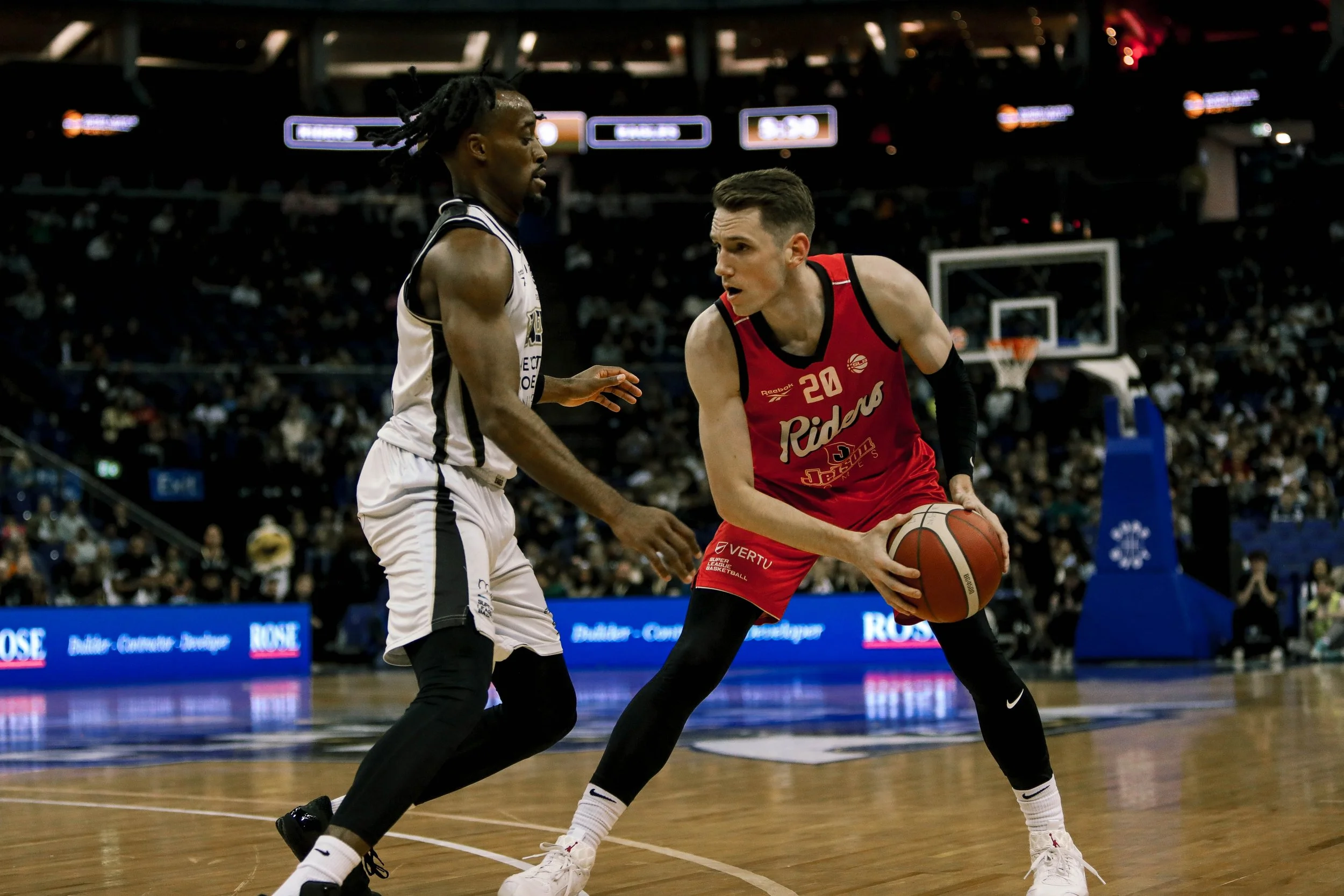 Two basketball players compete on the court during a game, with fans watching in the background.