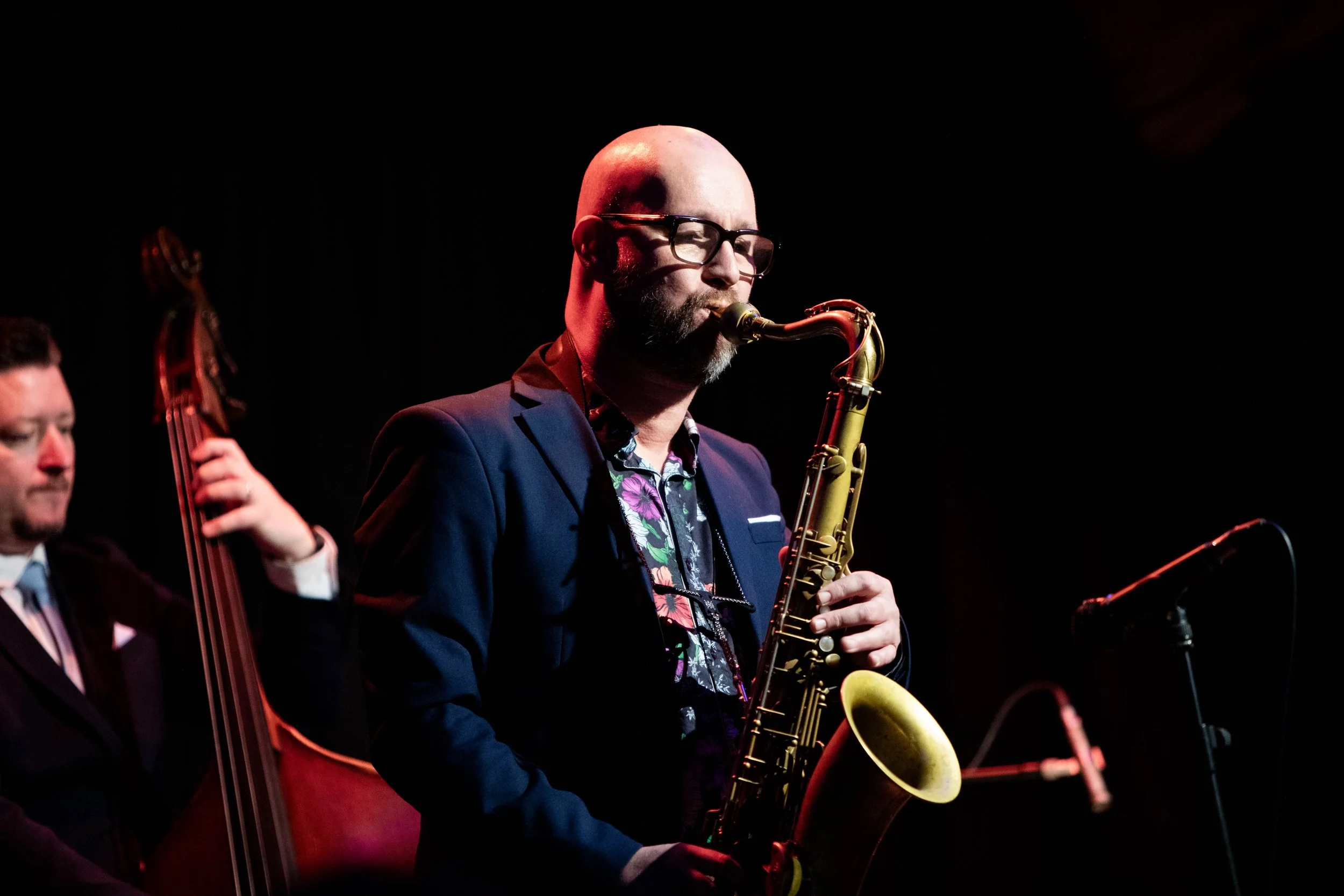 A man with glasses and a beard playing a saxophone on stage, with a microphone nearby, in a dimly lit setting.