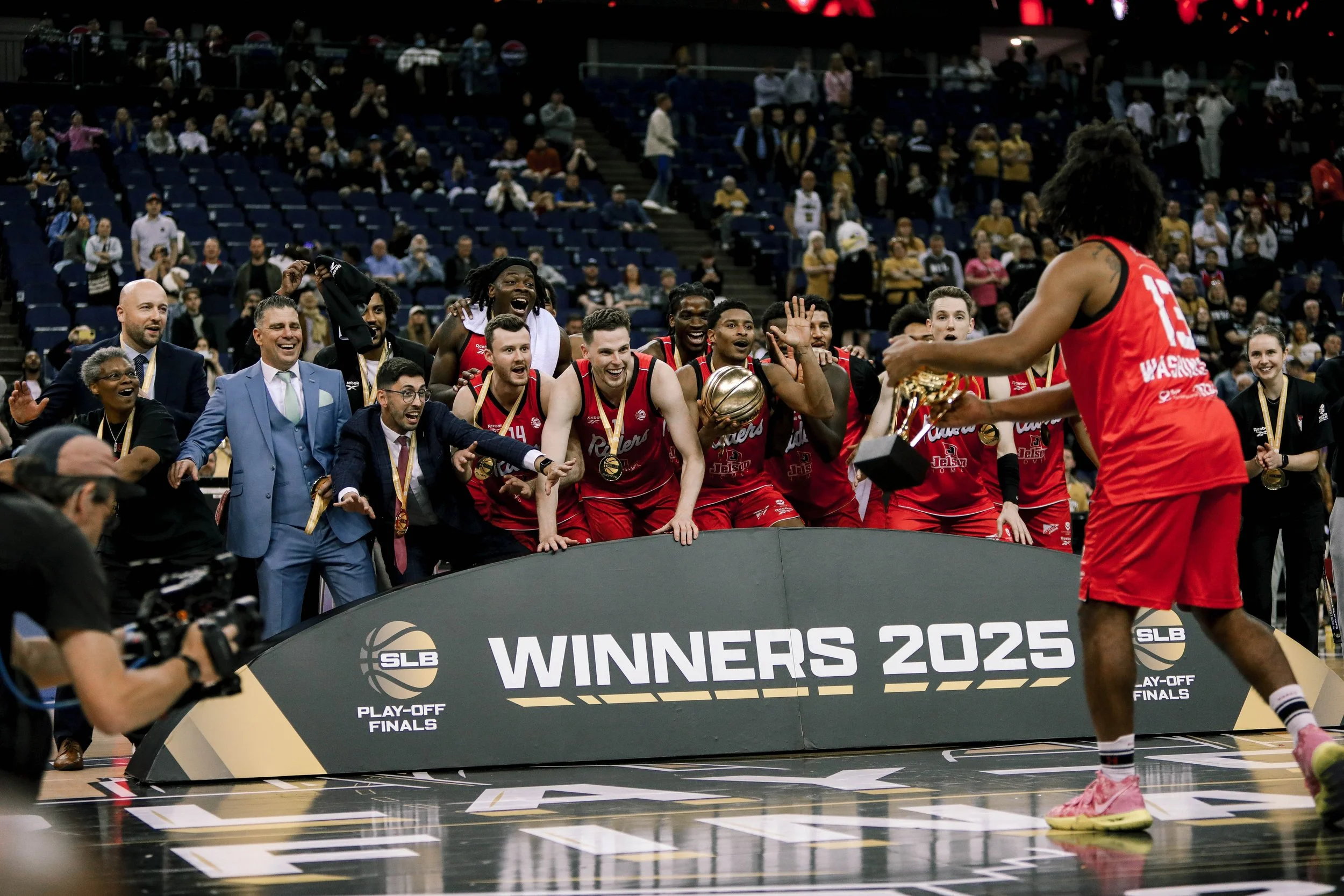 A basketball team celebrating their victory on the court, with medals around their necks, holding a trophy, and smiling, while a player in a red uniform approaches them, all in an arena with fans in the background.