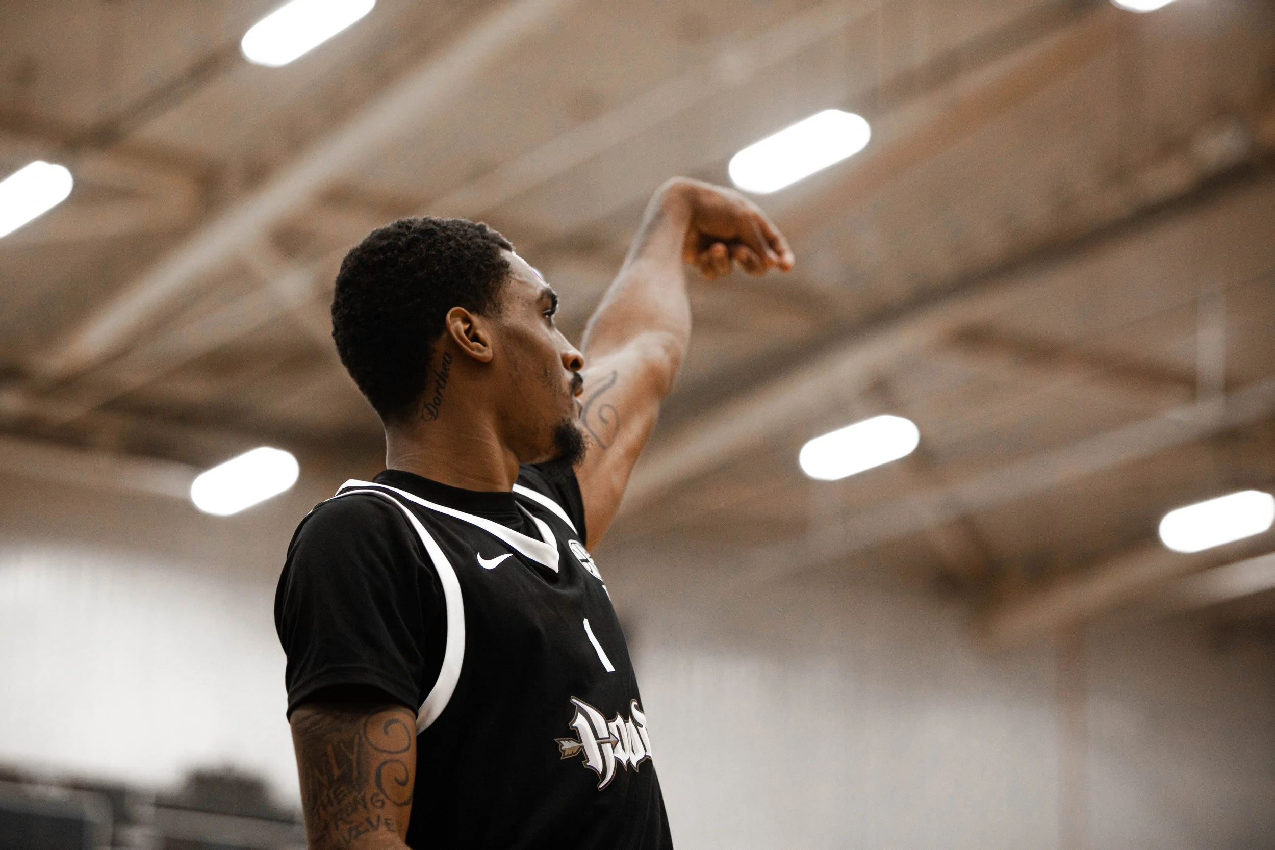 A male basketball player in a black jersey with white accents, standing in an indoor gymnasium, raising his left arm with his index finger pointing upward.