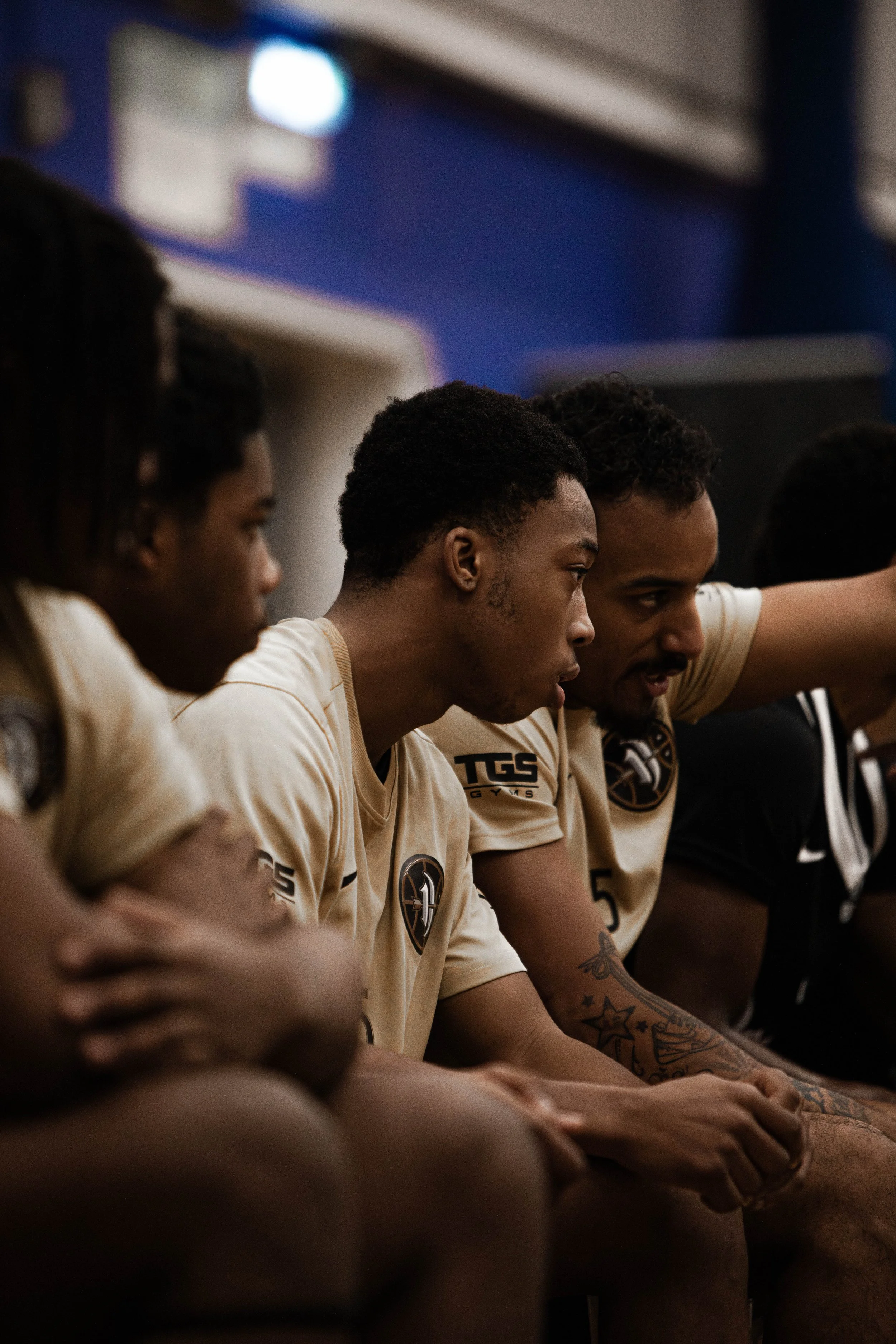 A group of young men in beige soccer jerseys sitting on a bench, some with visible tattoos, focused and engaged during a timeout or team huddle.