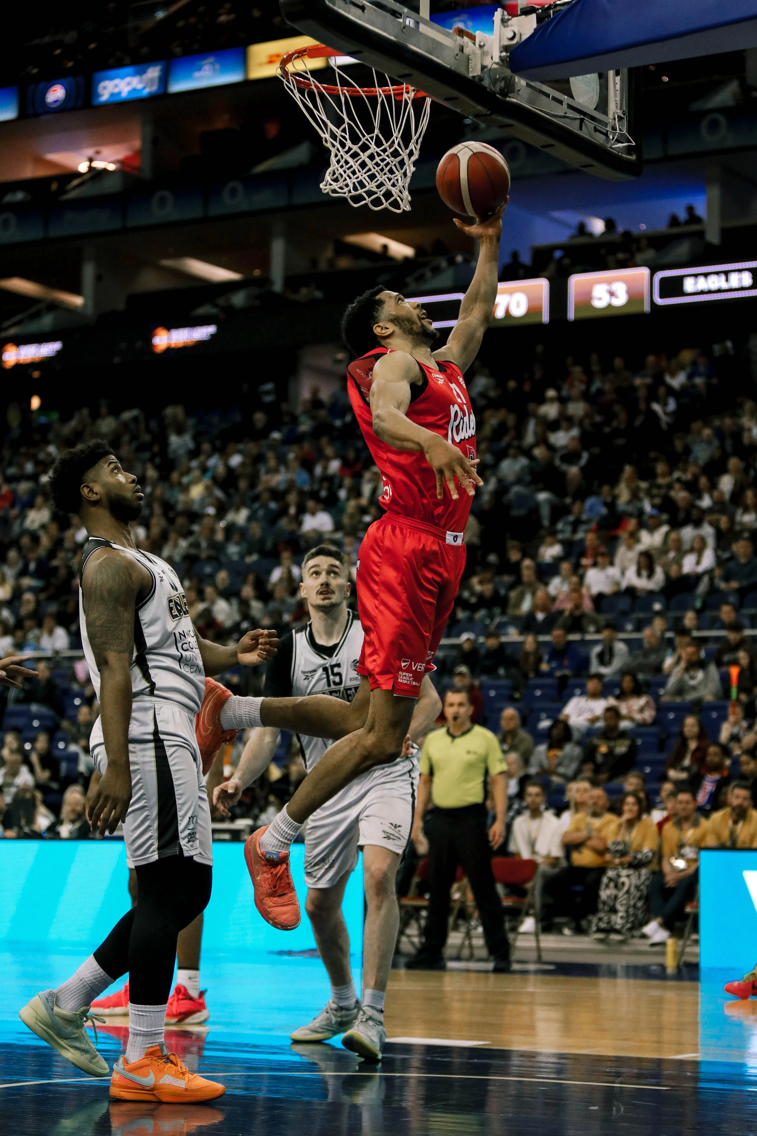 A basketball player in a red uniform is dunking the ball into the basket, while two players in white uniforms watch. The court is in an indoor arena with a crowd of spectators in the background.