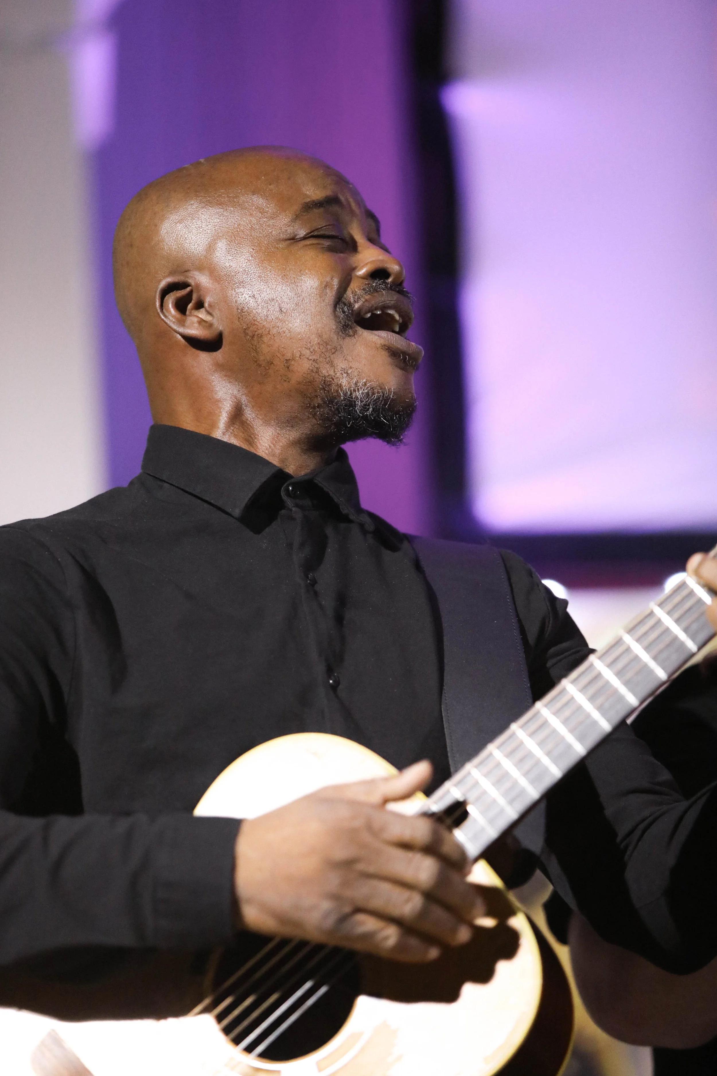 A man singing and playing an acoustic guitar, wearing a black shirt, with his eyes closed and an expressive face.