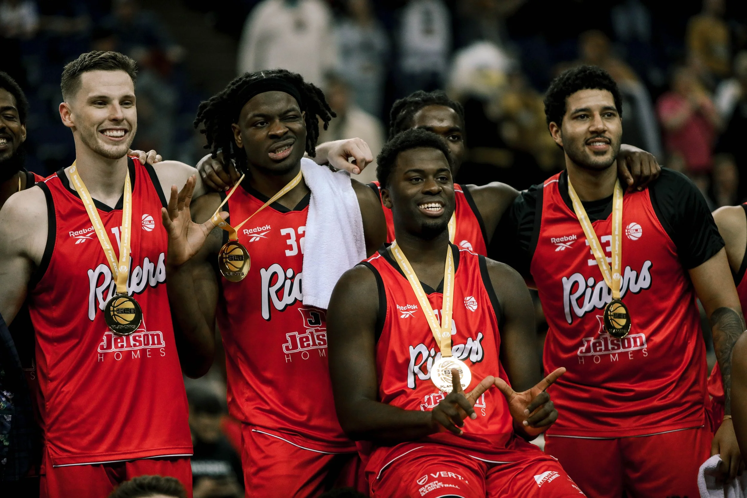 Group of basketball players in red jerseys celebrating with medals around their necks after a game, indoors in a stadium.