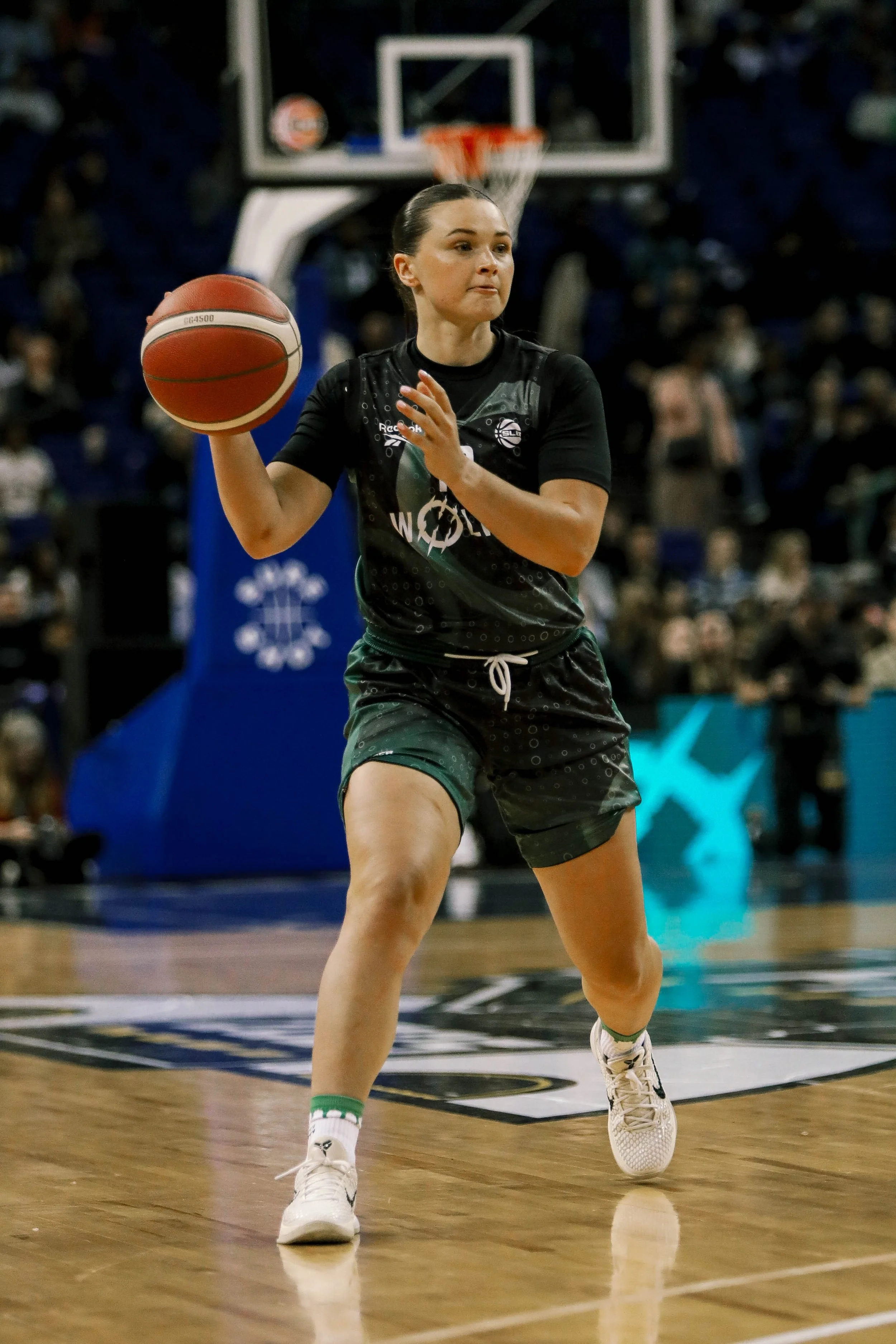 A woman playing basketball on an indoor court, preparing to shoot or pass the ball, with a basketball hoop in the background and spectators watching.