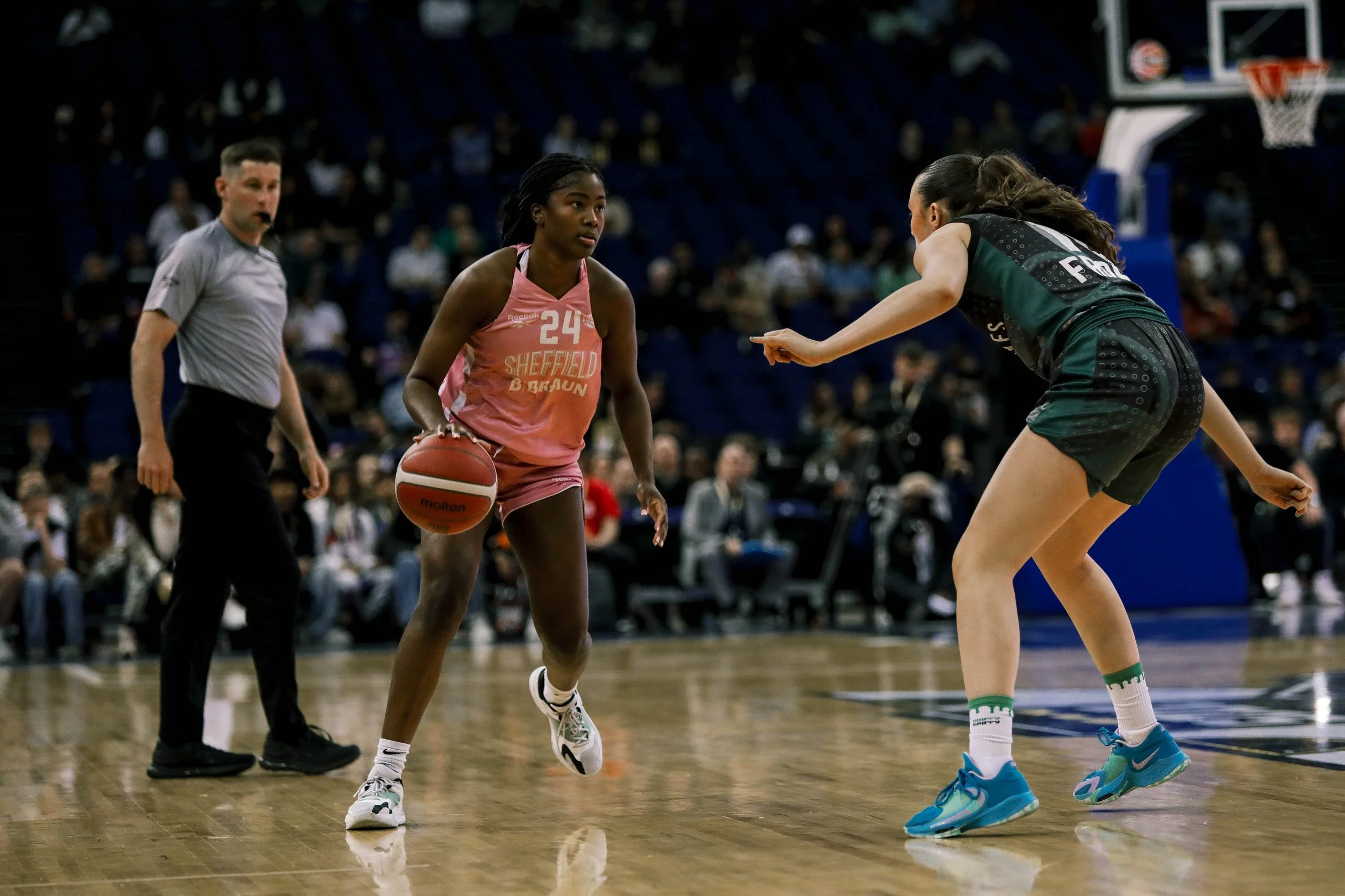 A women's basketball game with one player dribbling the ball while another defender guards her, a referee nearby, and spectators watching in the background.