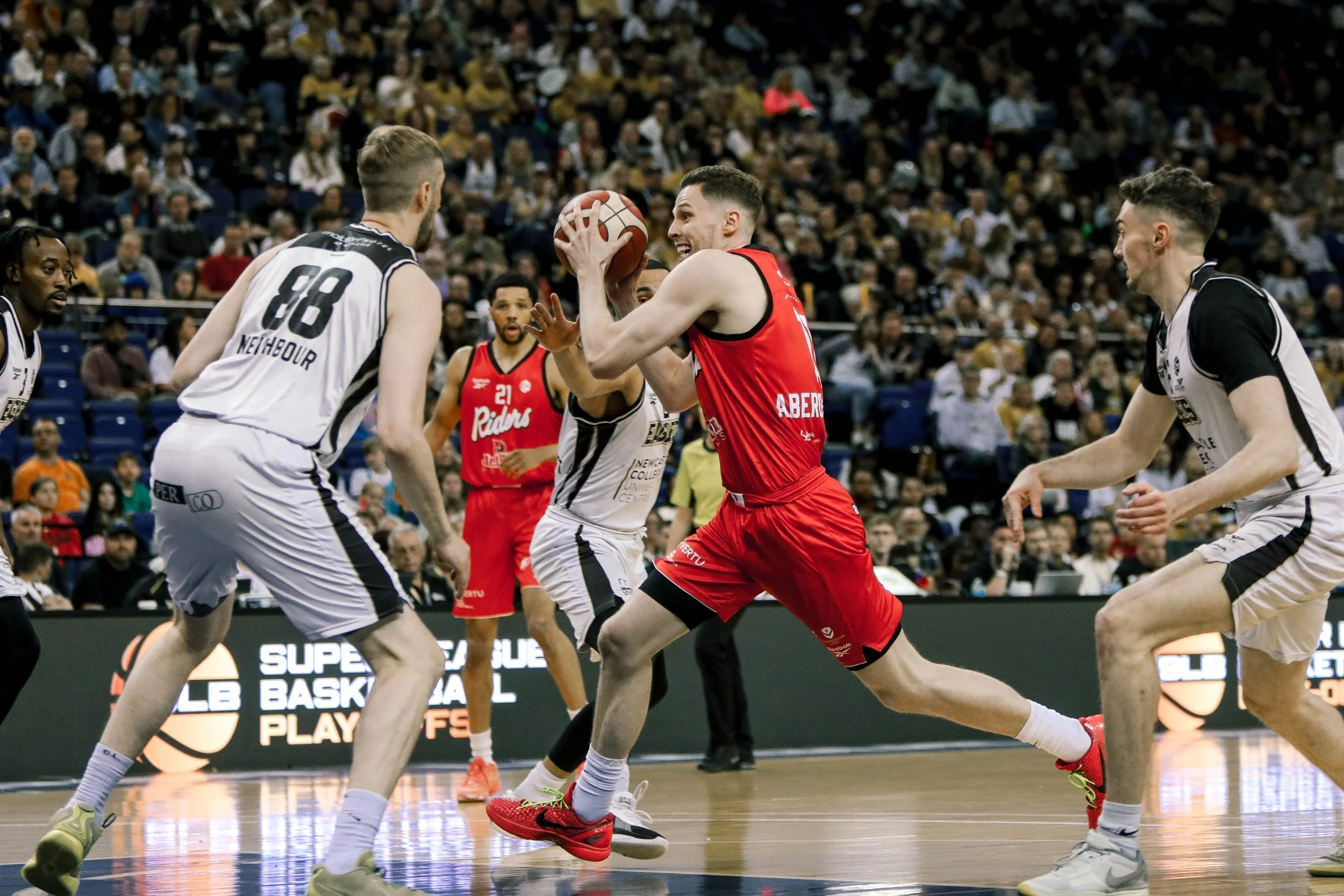 Basketball players in action during a game, with one player in a red uniform holding the ball and others in white and black uniforms defending, in a crowded indoor court setting.