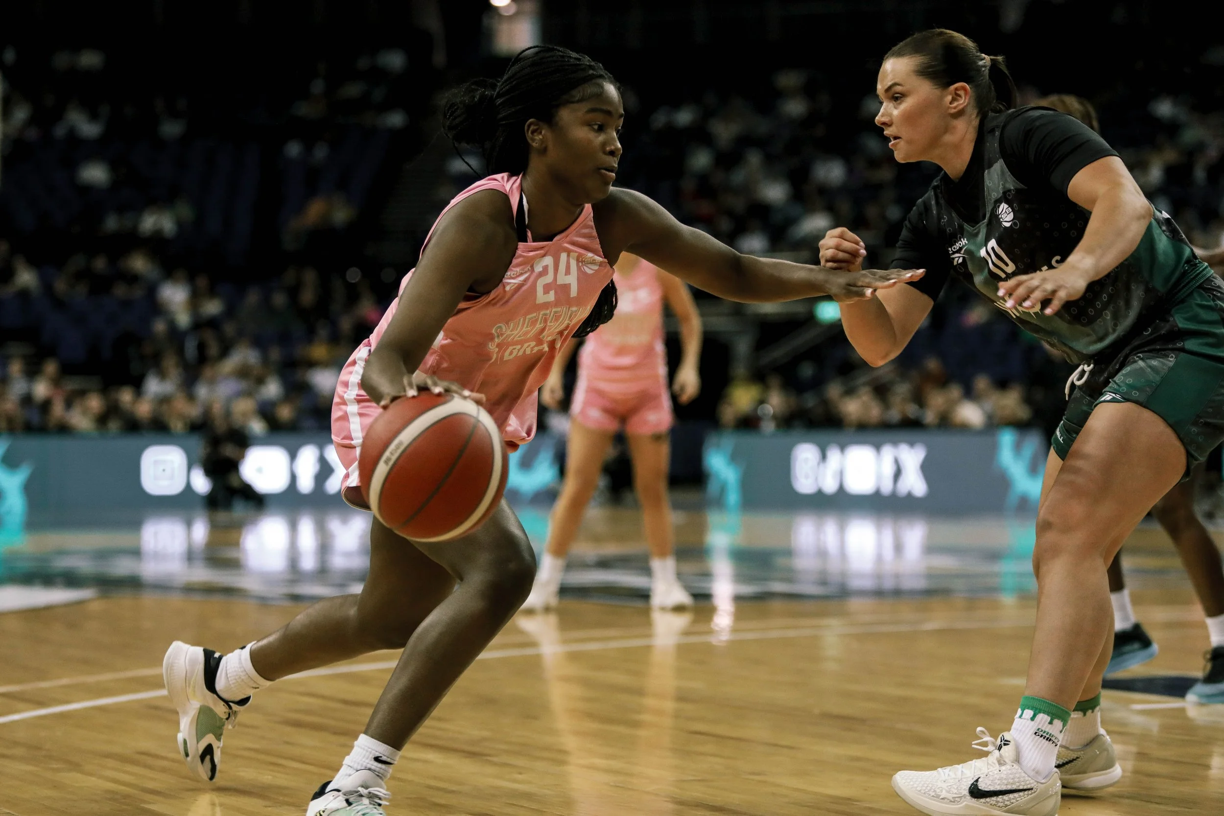 Two female basketball players are competing during a game, with one player dribbling and the other defending on a basketball court.